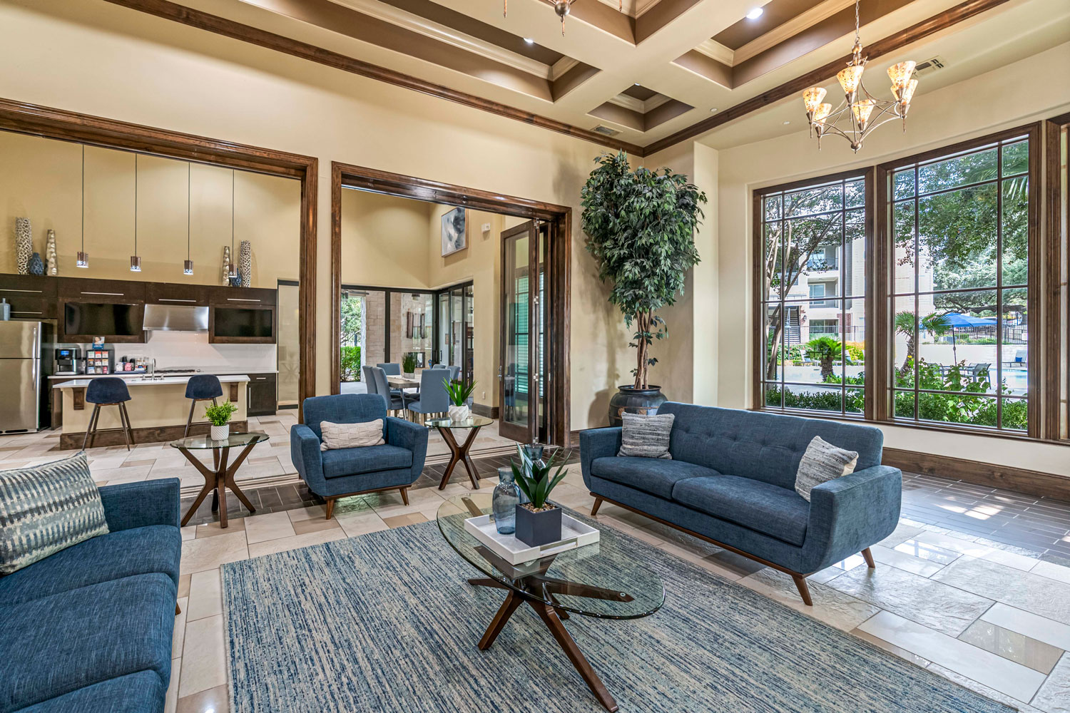 Spacious clubhouse lounge with blue sofas, glass coffee table, coffered ceiling, and open view to coffee bar and dining area