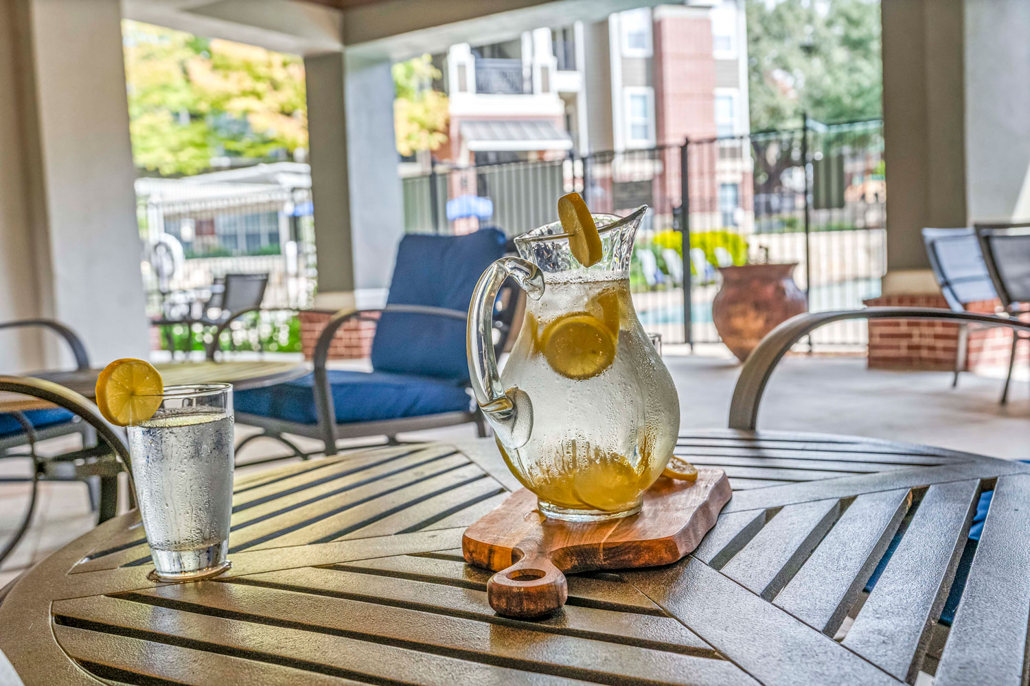 Lemon water pitcher and glass on patio table with cushioned outdoor lounge chairs and fenced swimming pool in background
