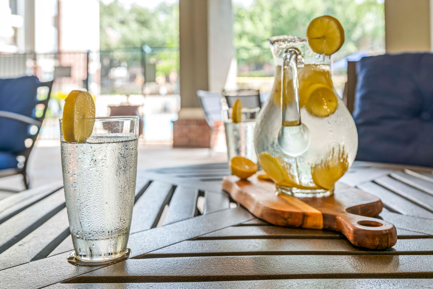 Glass of lemon water beside pitcher with citrus slices on outdoor table under covered patio seating area near community pool