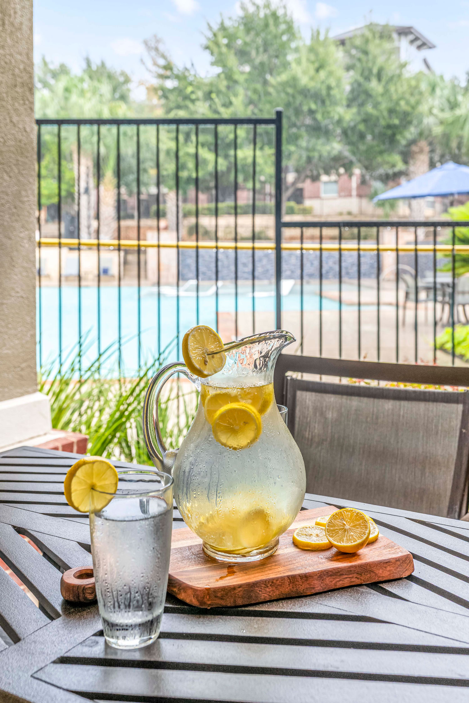 Lemon water pitcher and drinking glass on patio table overlooking resort style pool area behind black metal safety fence