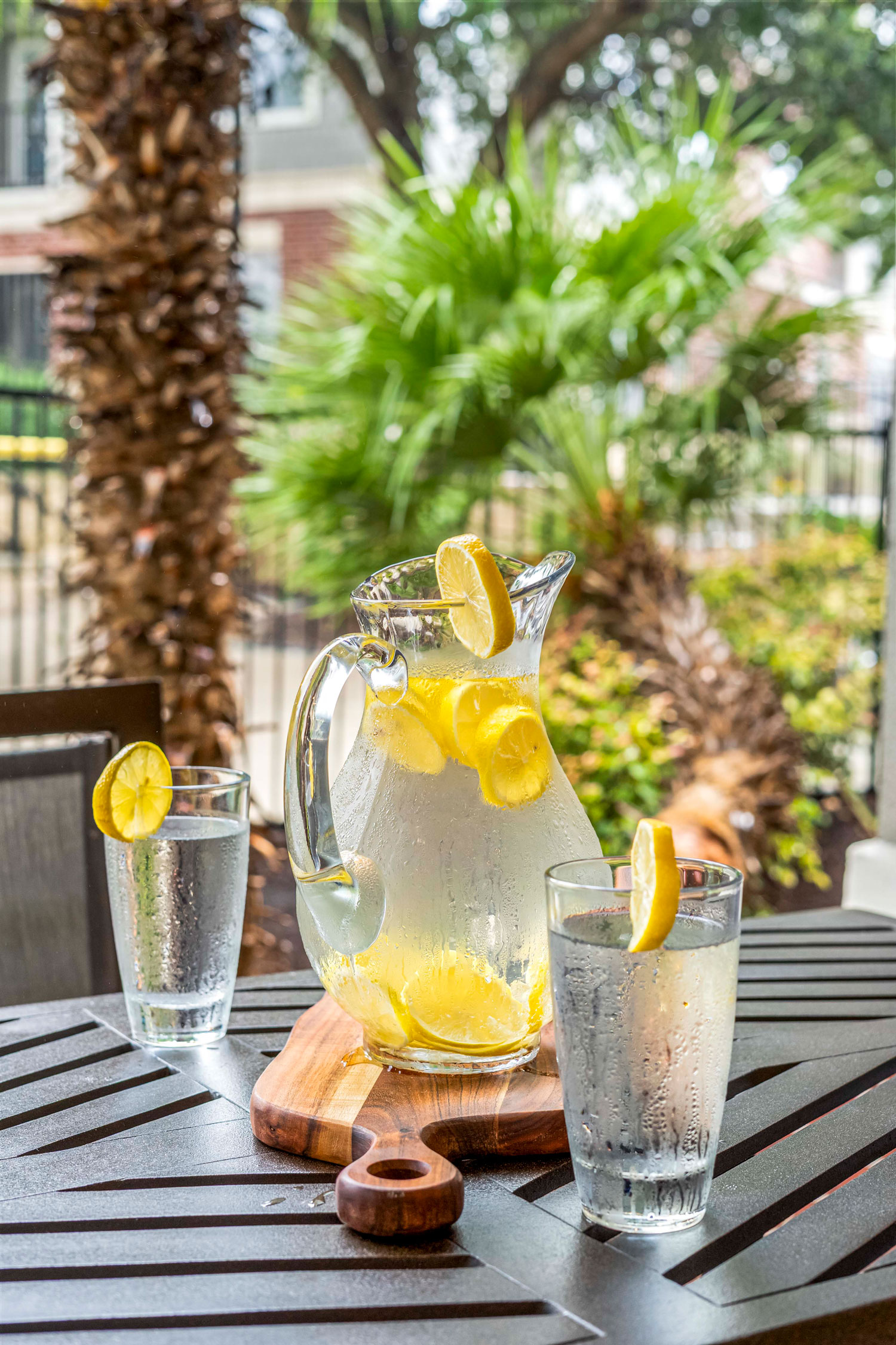 Glass pitcher of lemon water and two drinking glasses on outdoor patio table with palm trees and landscaped apartment courtyard