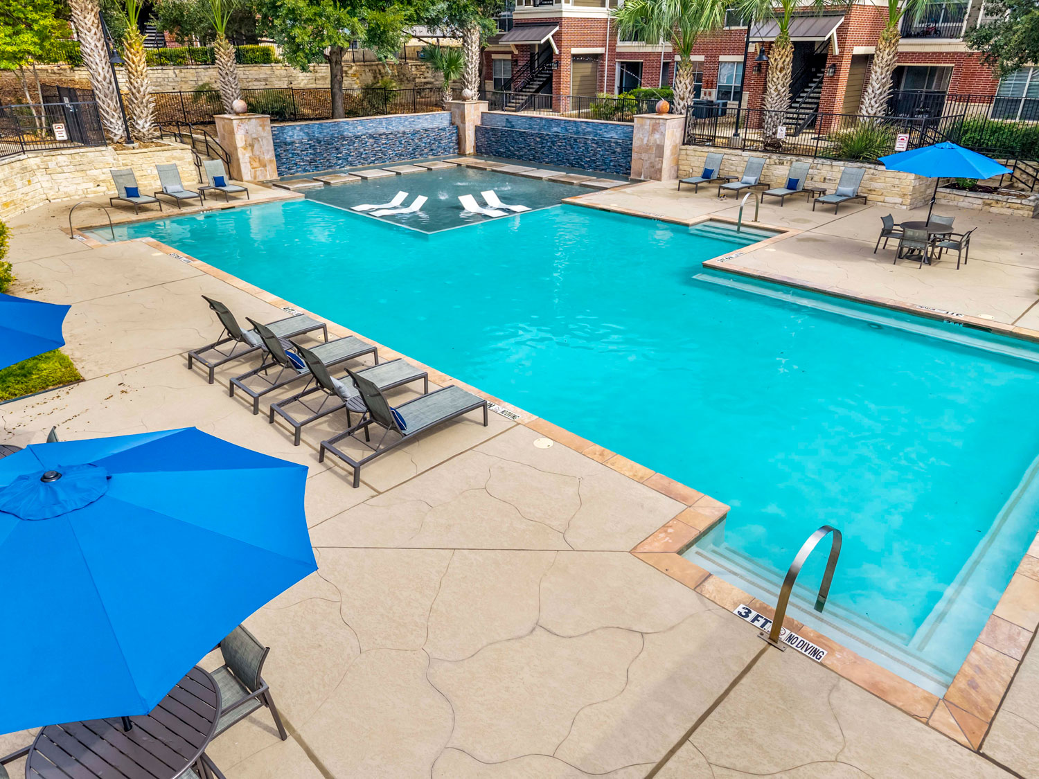 Resort-style community pool with blue lounge chairs, patio umbrellas, a tanning ledge, and a decorative mosaic water wall