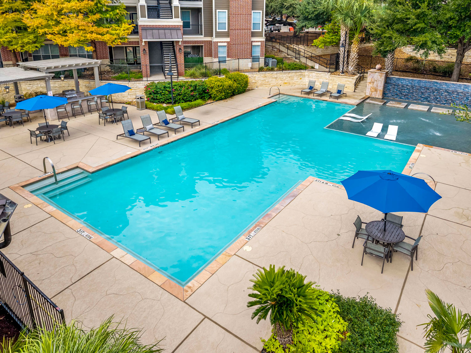 Elevated view of a resort-style pool with blue umbrellas, lounge chairs, a tanning ledge, and a mosaic tile water feature