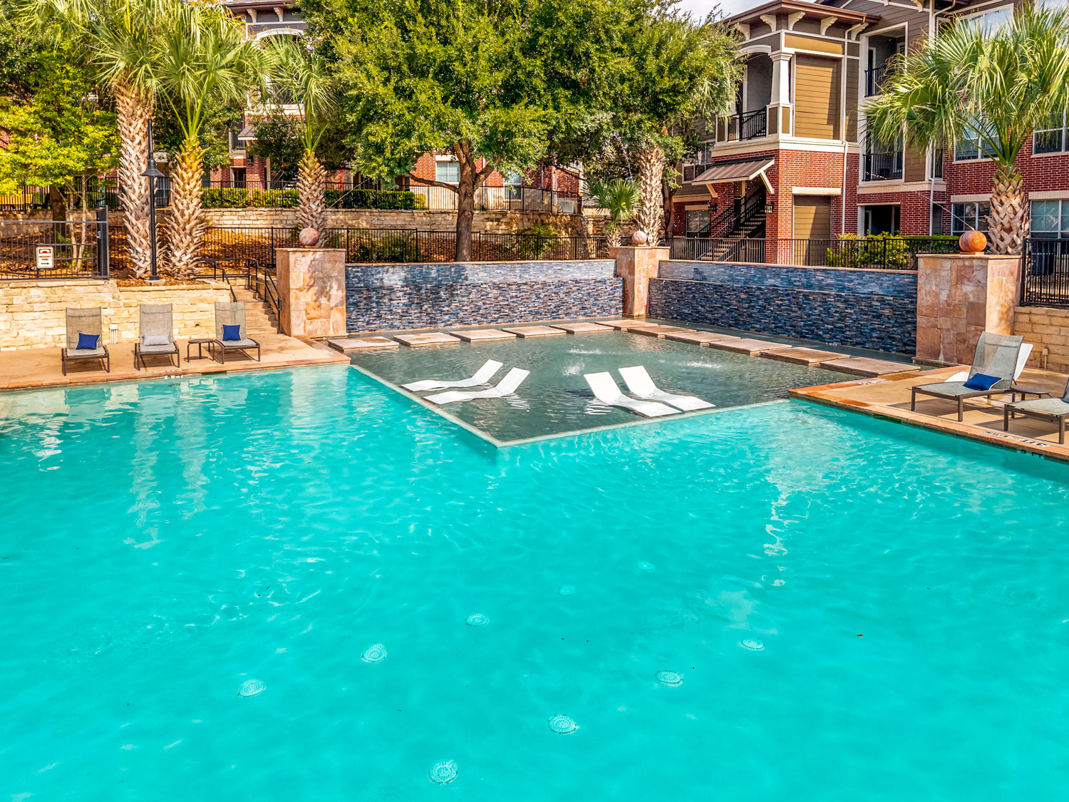 Resort style swimming pool with tanning ledge loungers, bubbling water features, palm trees, and apartment buildings nearby
