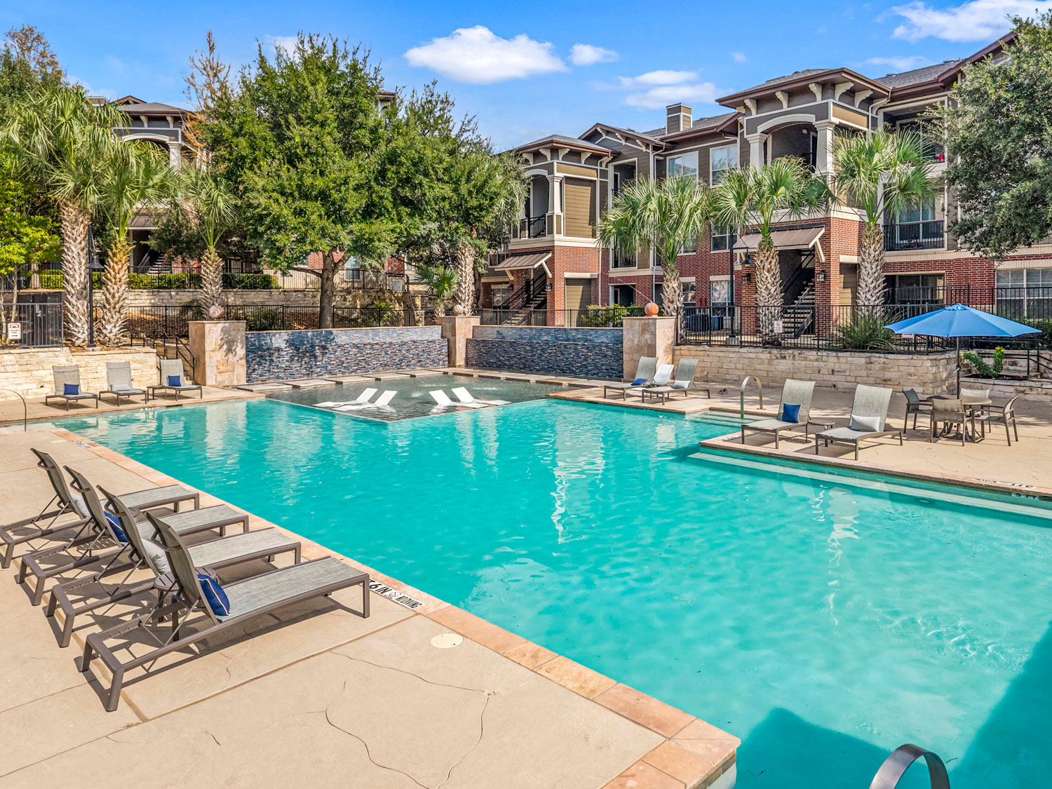 Resort style pool deck with lounge chairs, blue umbrellas, shaded pergola seating, and nearby apartment buildings