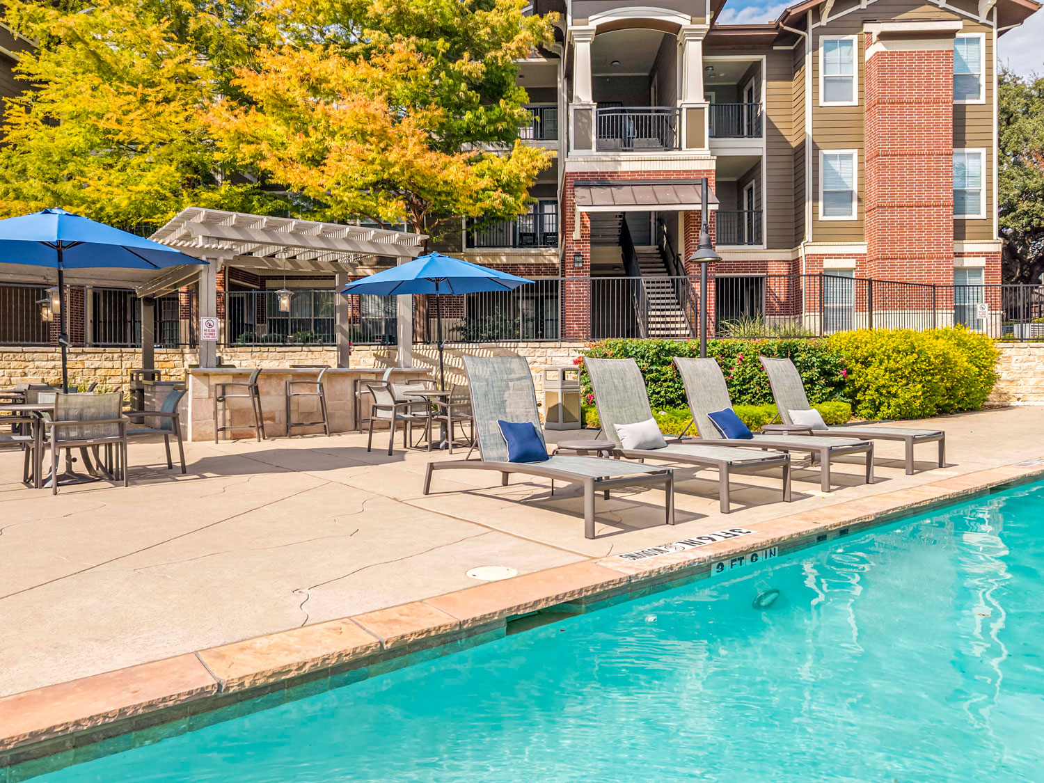 Large resort style swimming pool with lounge chairs, tanning ledge seating, palm trees, and surrounding apartment buildings