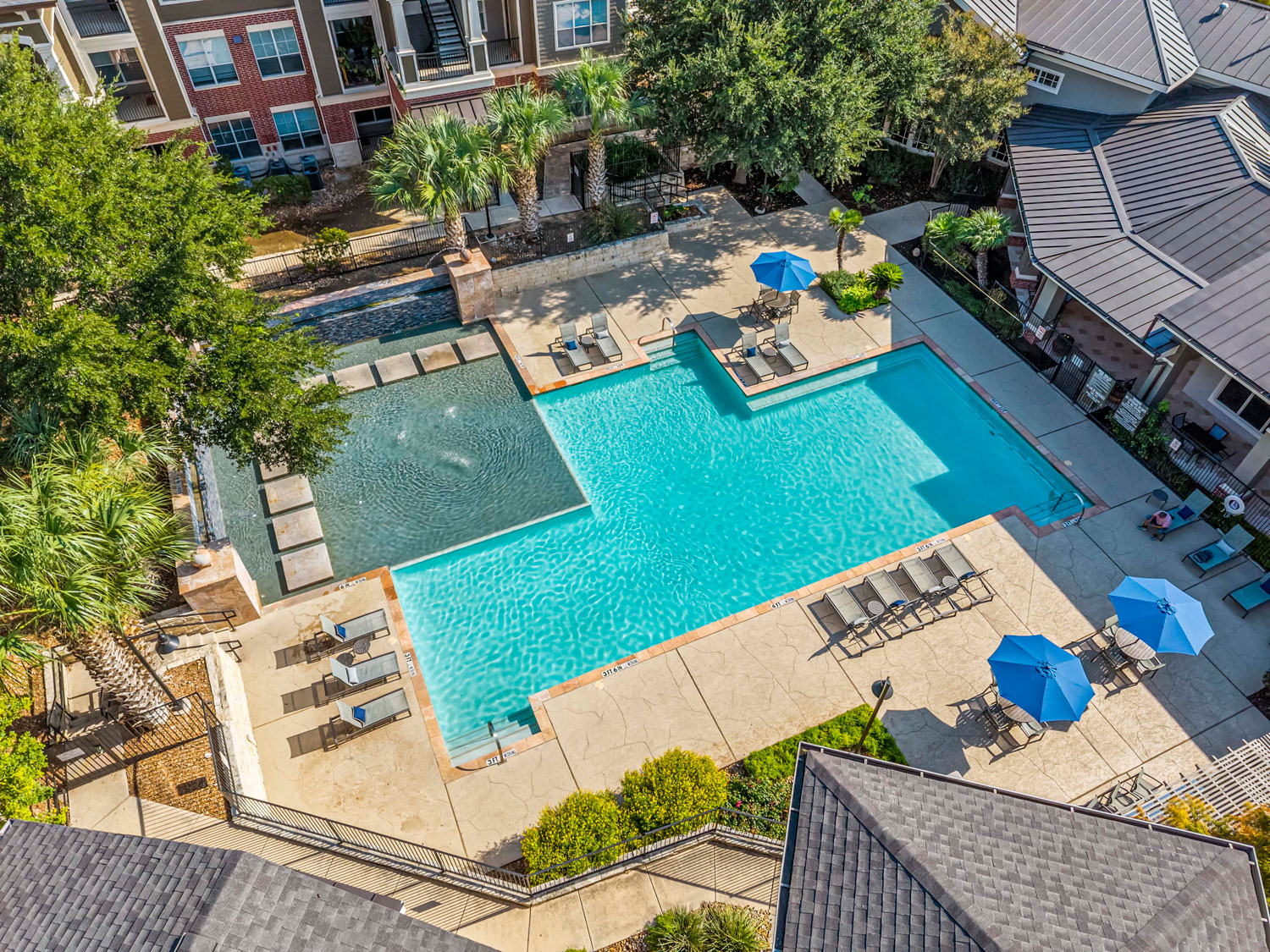 Drone aerial view of a resort-style pool and spa with lounge chairs, blue umbrellas, and palm trees surrounded by apartments