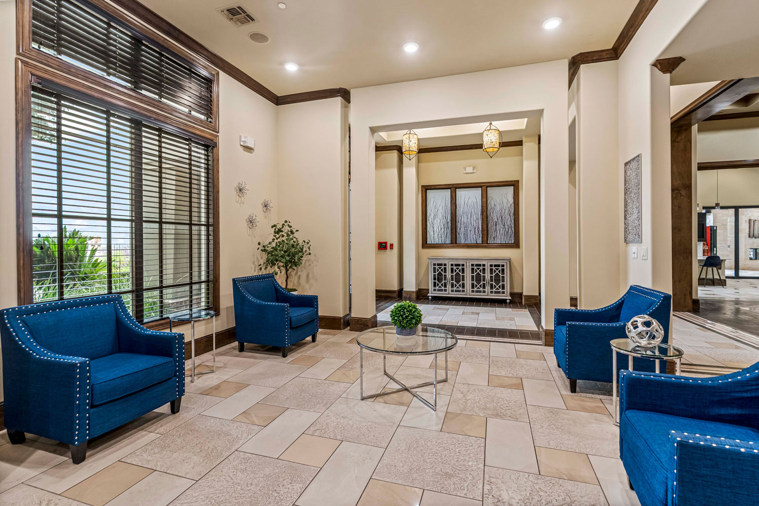 Apartment clubhouse lobby with blue lounge chairs, glass coffee table, large windows, and decorative lighting fixtures