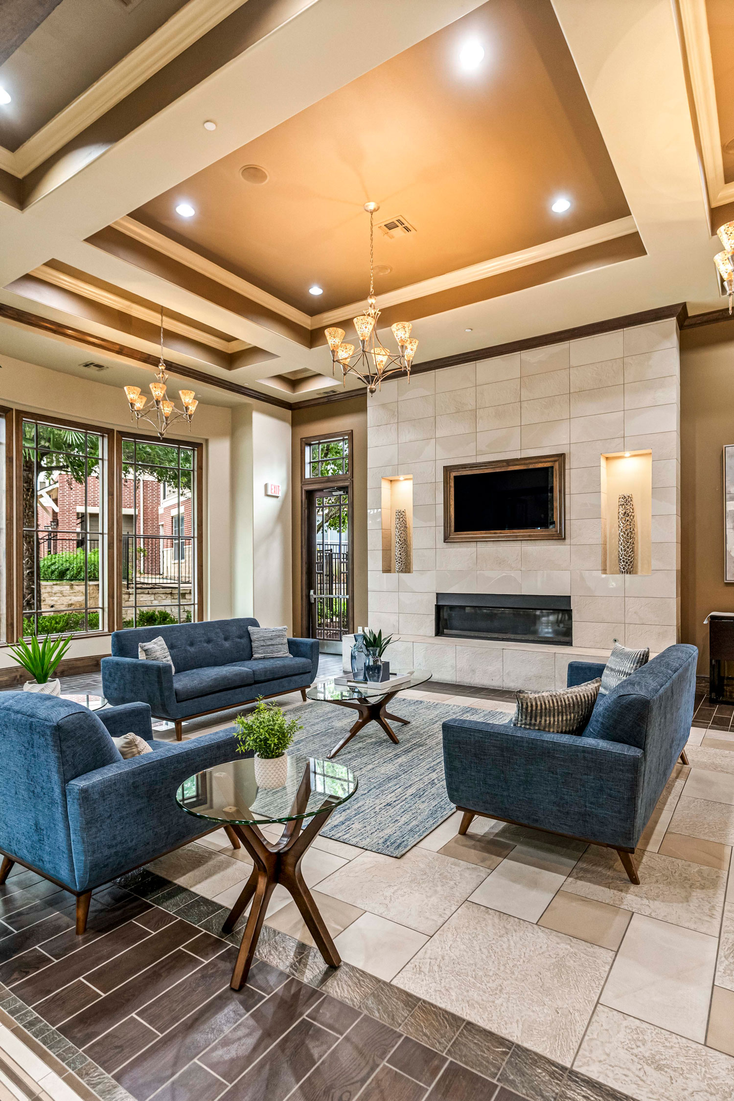 Resident lounge with blue seating, glass coffee tables, decorative fireplace wall, and high coffered ceiling with warm pendant lighting
