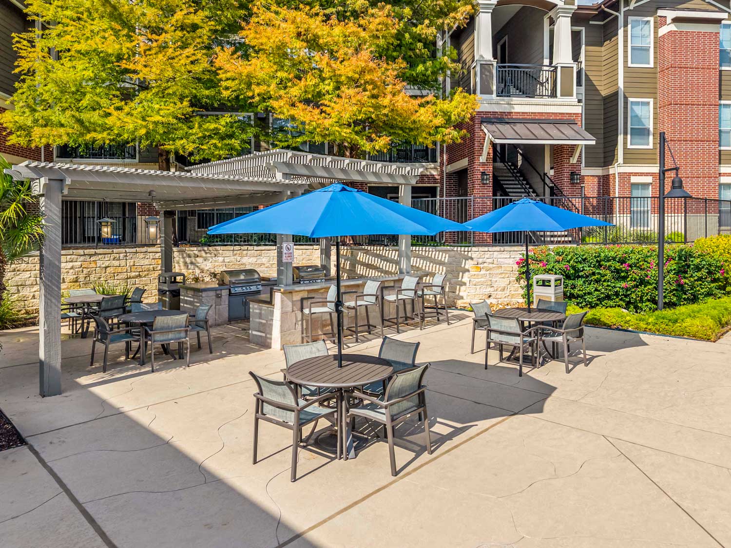 Outdoor grilling pavilion with stainless grills, bar seating, dining tables, blue umbrellas, and landscaped courtyard patio