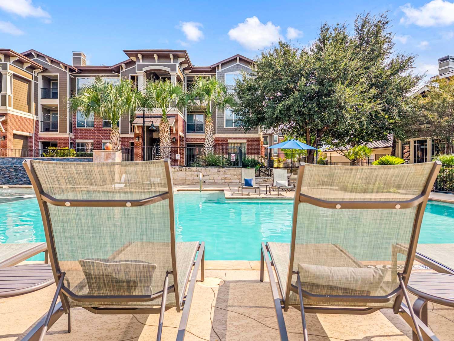Poolside lounge chairs facing resort style swimming pool with palm trees, landscaped courtyard, and apartment buildings in background