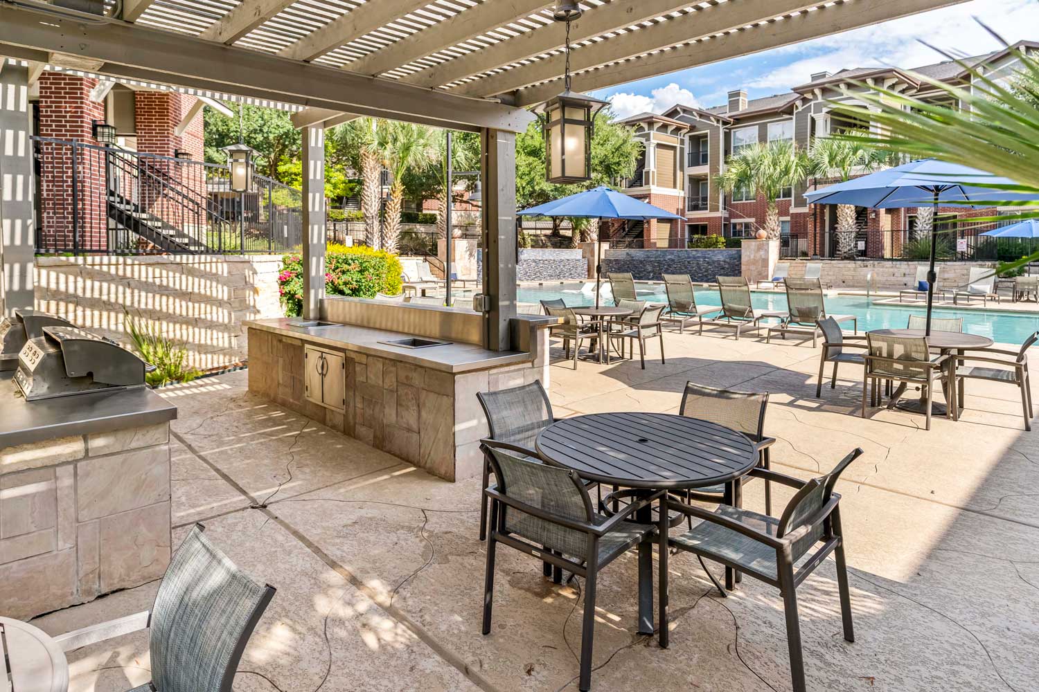 Covered outdoor grilling pavilion with pergola roof, stainless grill, prep counter, and poolside patio seating area