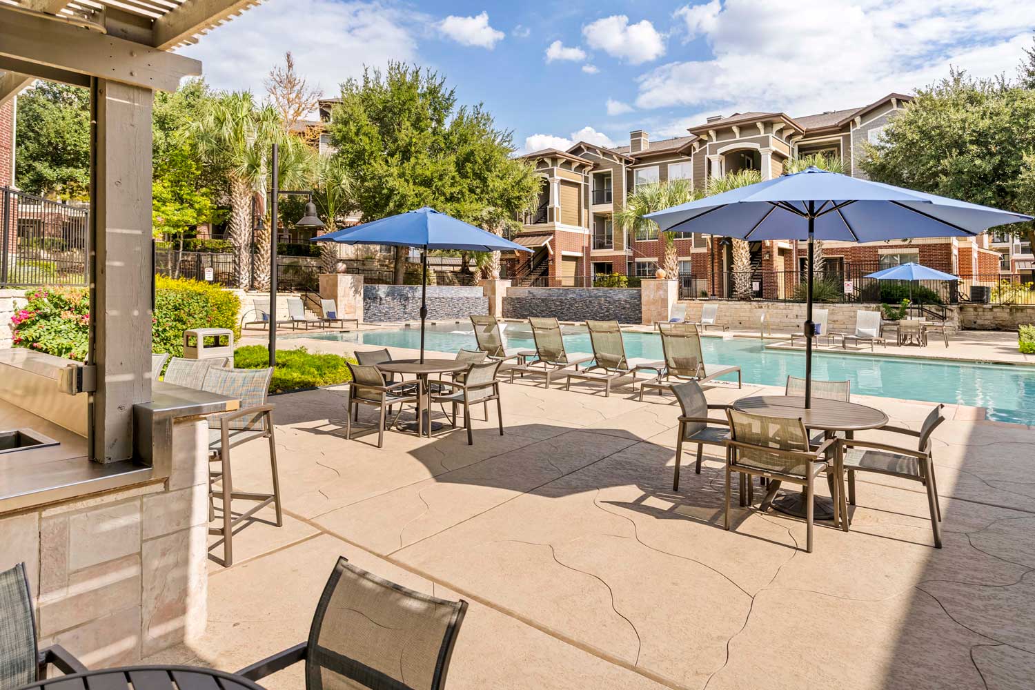 Poolside patio with dining tables, blue umbrellas, lounge chairs, and landscaped resort style swimming pool courtyard