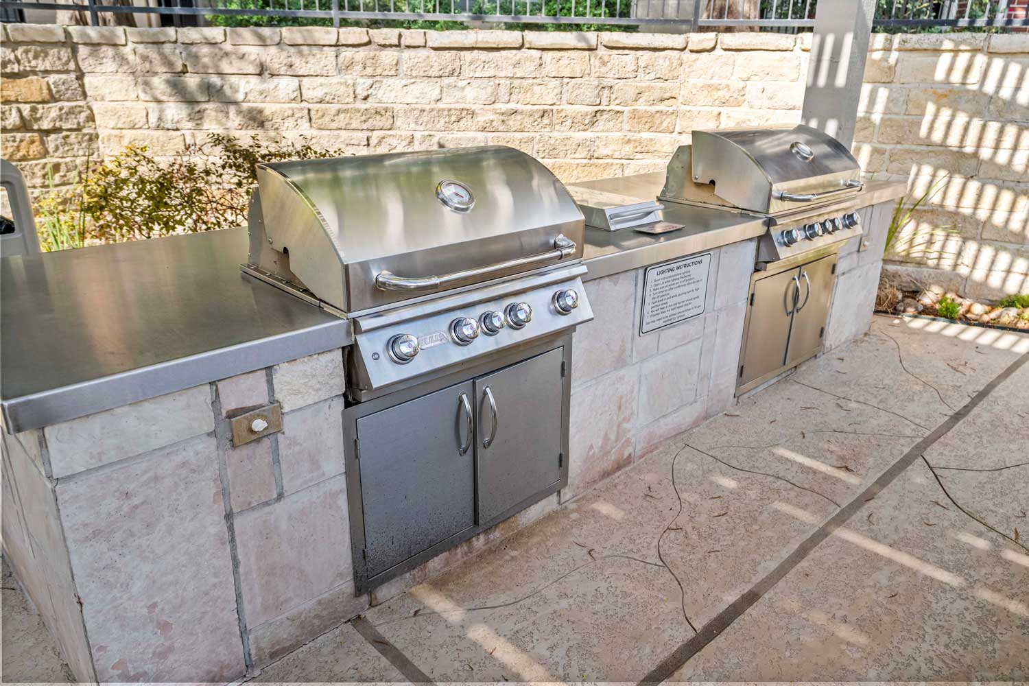 Outdoor grilling station with stainless gas grills, stone counter surround, and pergola shade structure in landscaped courtyard
