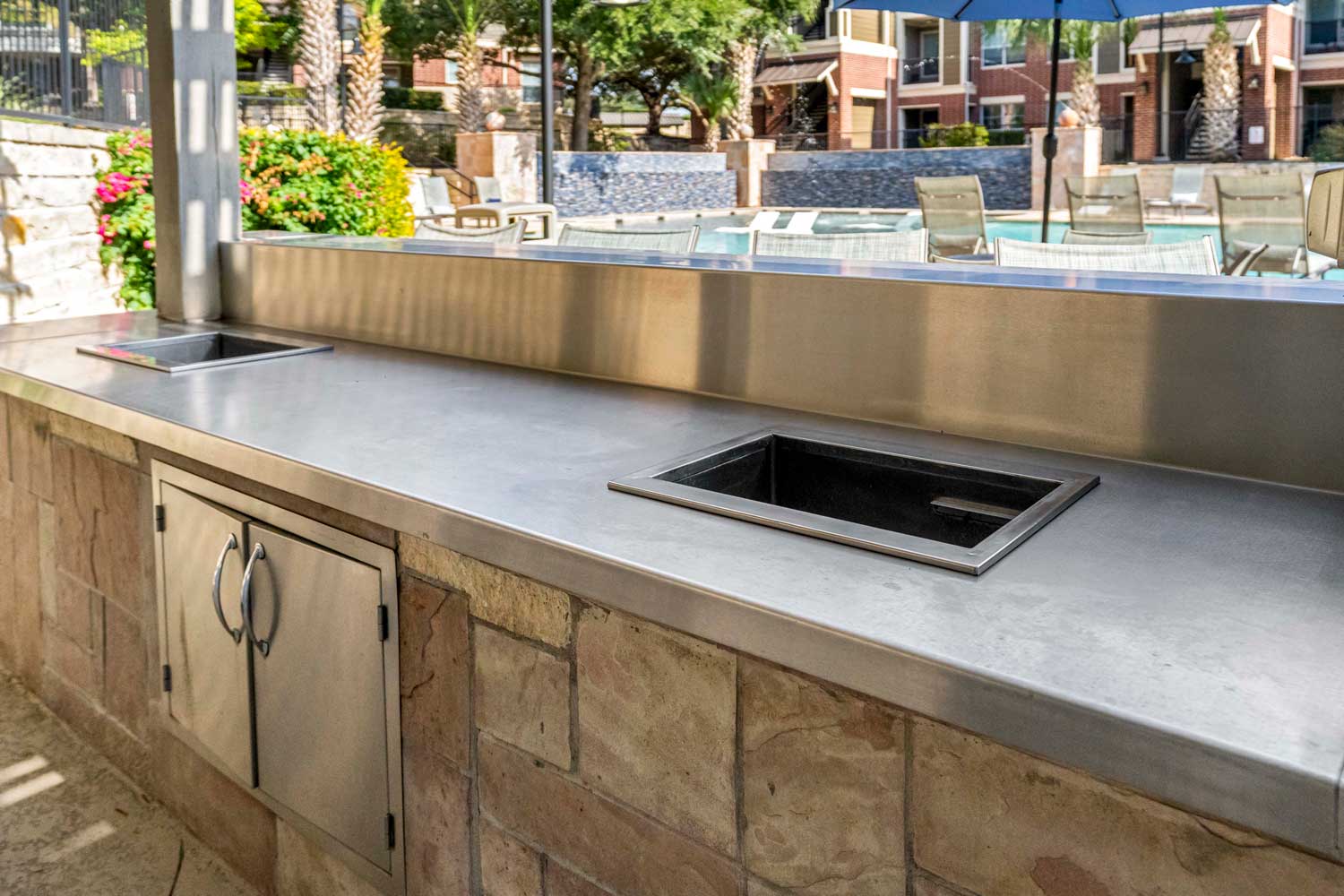 Outdoor grilling pavilion counter with stainless prep surface, built in sinks, and resort style pool courtyard in background