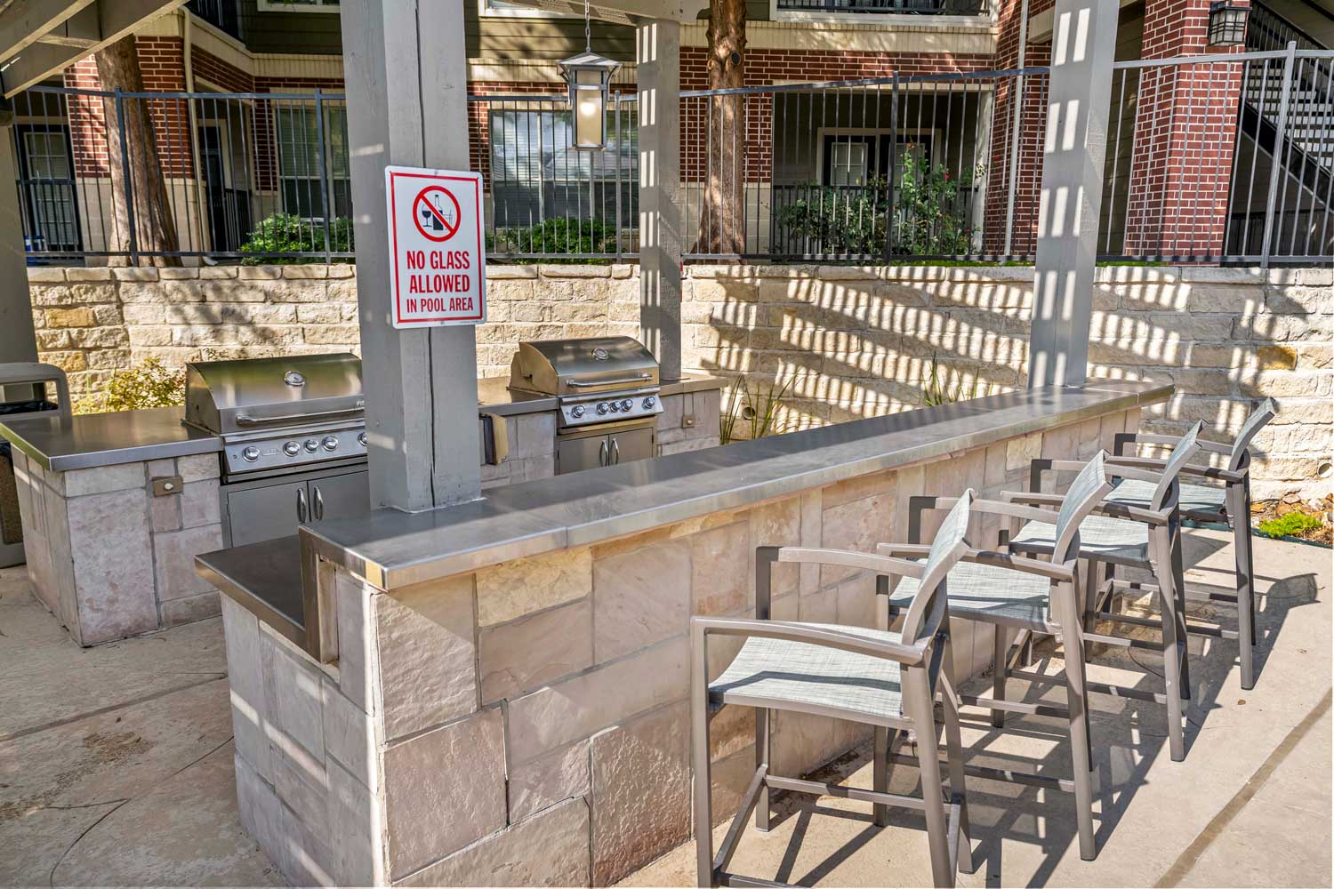 Outdoor grilling station with stainless grills, stone counter bar seating, pergola shade structure, and community courtyard backdrop