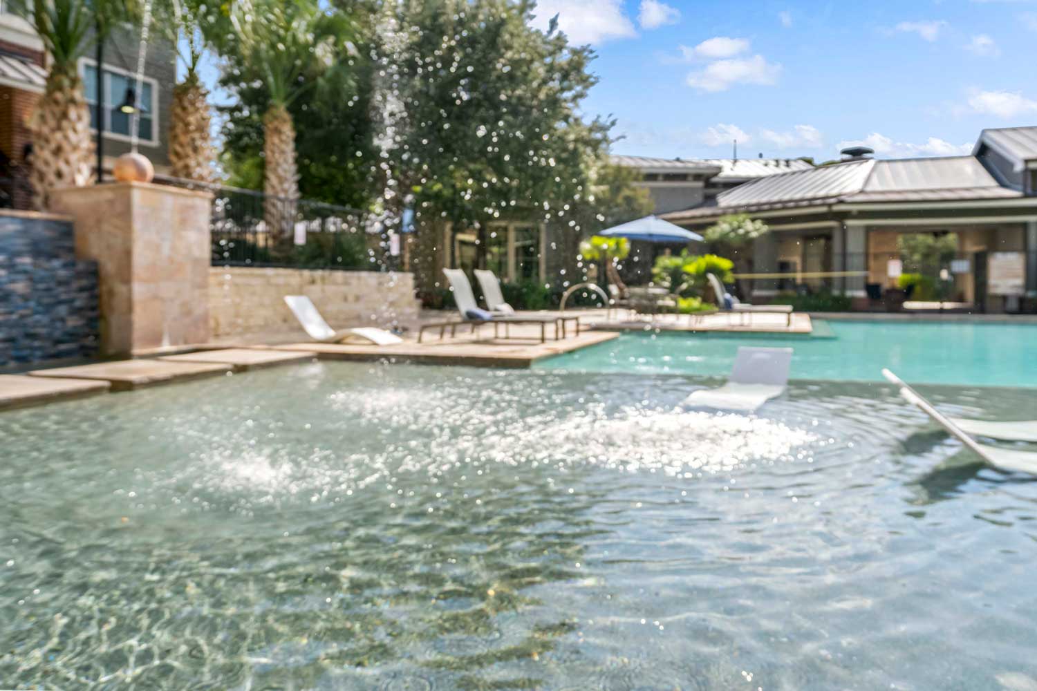 Shallow tanning ledge with in-pool lounge chairs, bubbling water feature, and resort style swimming pool in landscaped courtyard