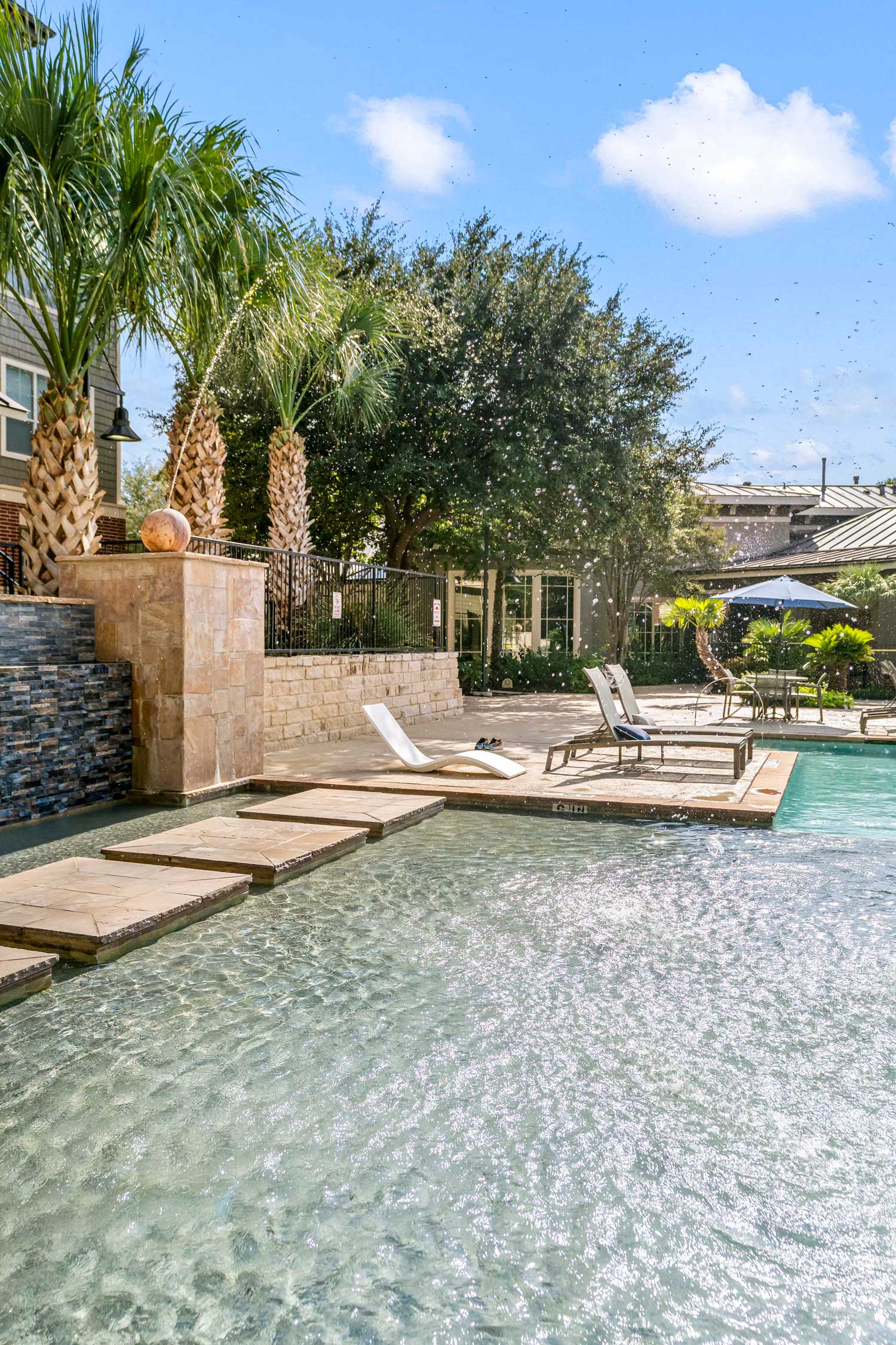Outdoor pool area with shallow water feature, sun loungers, palm trees, and landscaped courtyard seating areas