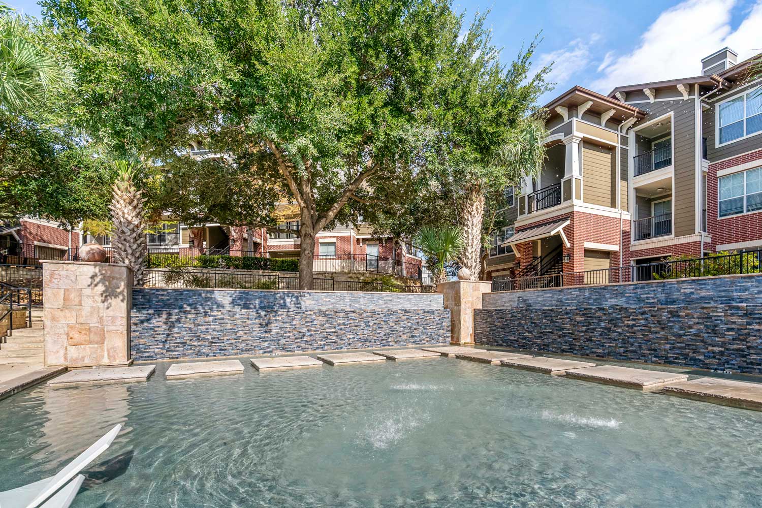 Pool courtyard with shallow splash area, decorative stone wall, palm trees, and nearby apartment community buildings