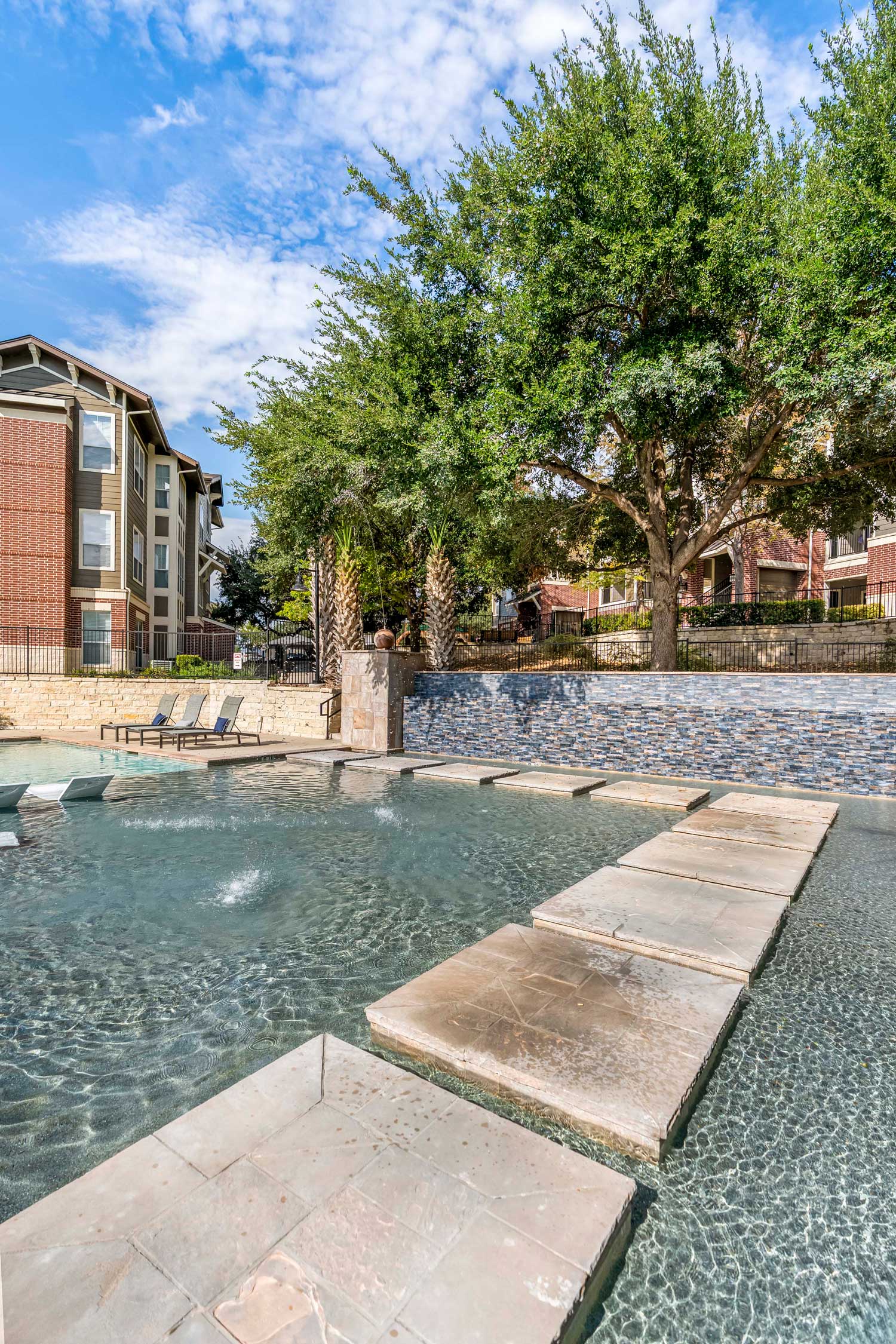 Resort style swimming pool with shallow water feature, stepping stones, stone wall accents, and surrounding apartment buildings