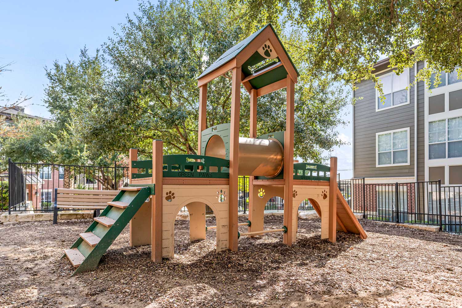 Outdoor pet play area with wooden agility structure, mulch ground cover, shade trees, fencing, and nearby apartment homes