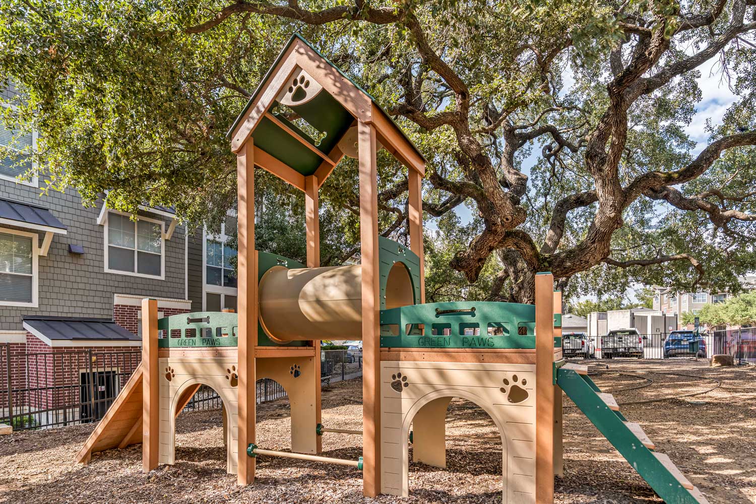 Pet play structure with ramps and tunnel features in shaded outdoor dog park surrounded by mature trees and apartment homes