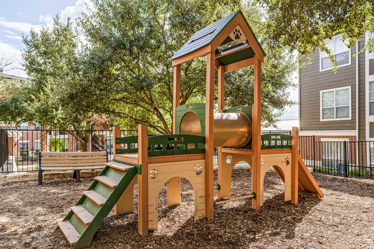 Pet-friendly outdoor play area with climbing structure, mulch ground cover, shaded trees, and nearby apartment buildings