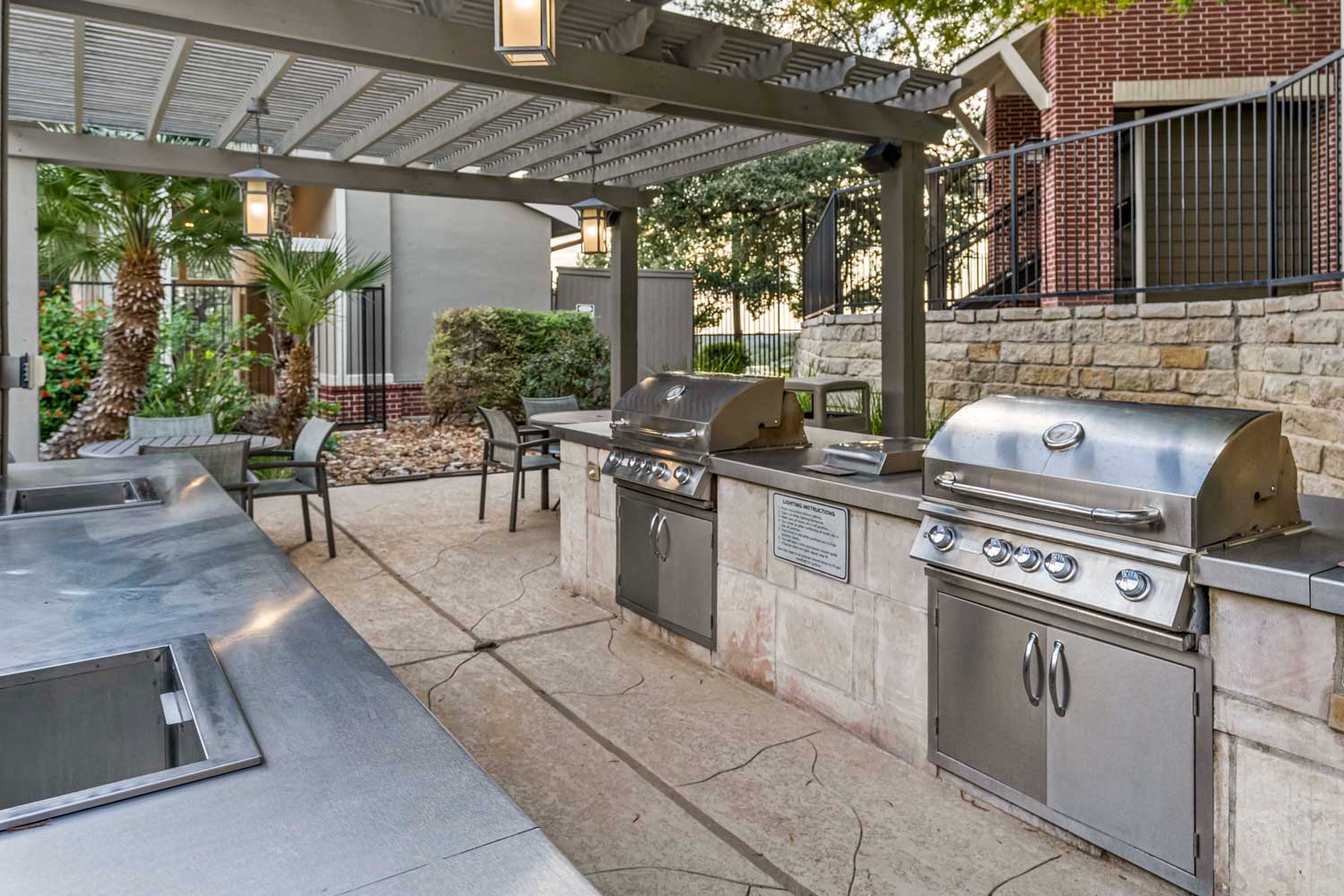 Covered outdoor grilling pavilion with stainless steel grills, prep counters, dining tables, and landscaped courtyard seating area