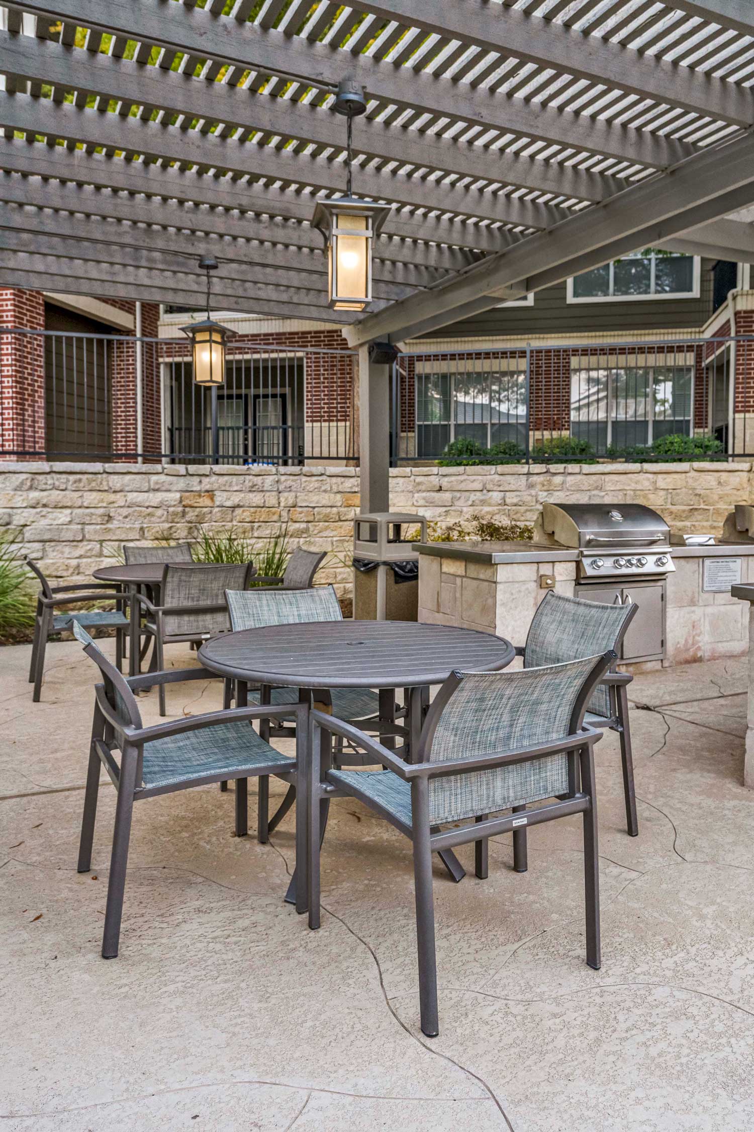 Outdoor grilling pavilion with pergola roof, hanging lantern lights, stainless grill, and patio table seating beside landscaped courtyard