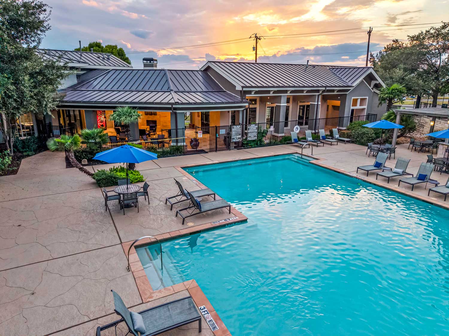 Resort style swimming pool with in-pool loungers, patio seating, palm trees, and illuminated clubhouse at sunset