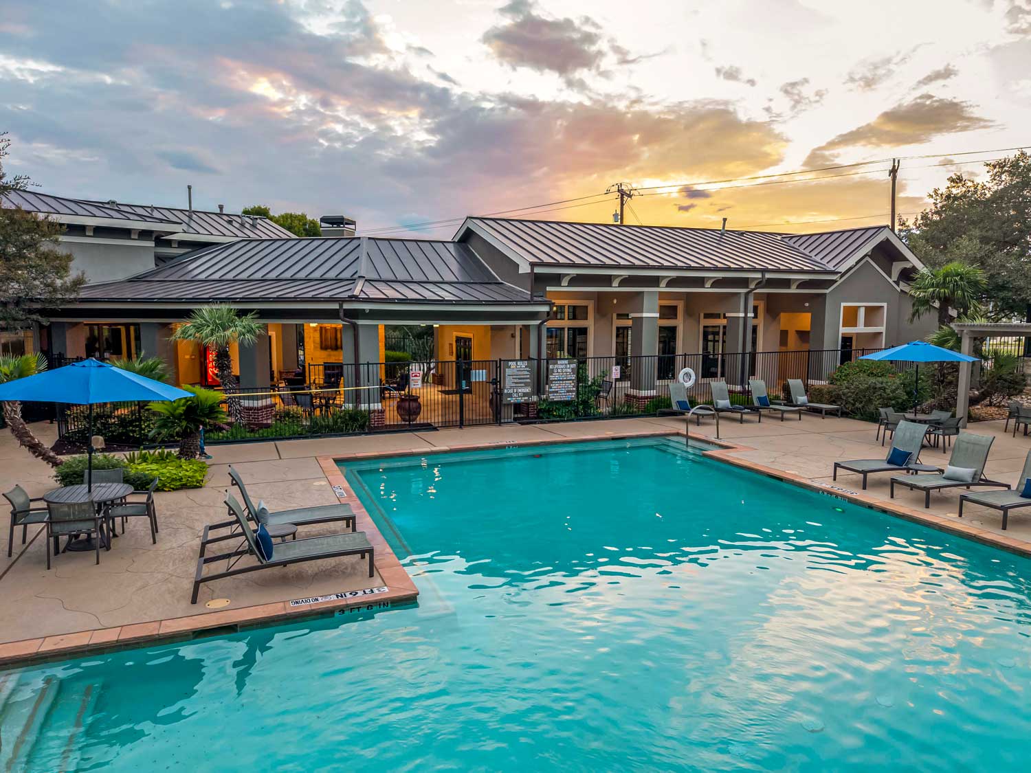 Resort style swimming pool with lounge chairs, patio tables, blue umbrellas, and clubhouse building at sunset