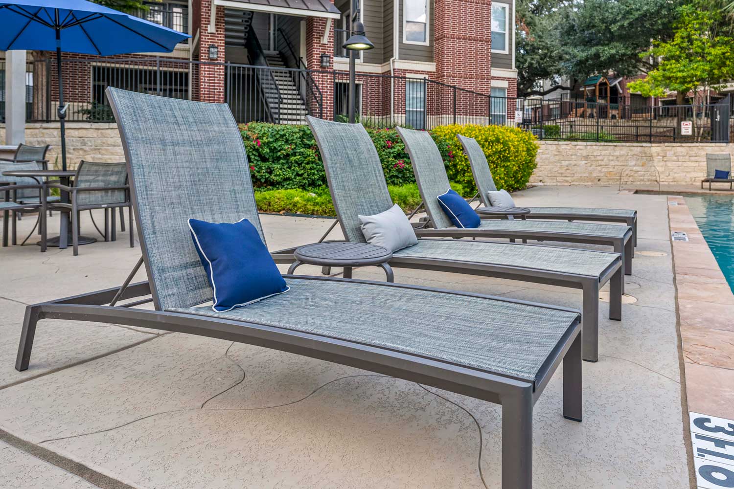 Poolside lounge chairs with cushions beside resort style swimming pool, patio tables, umbrellas, and landscaped courtyard