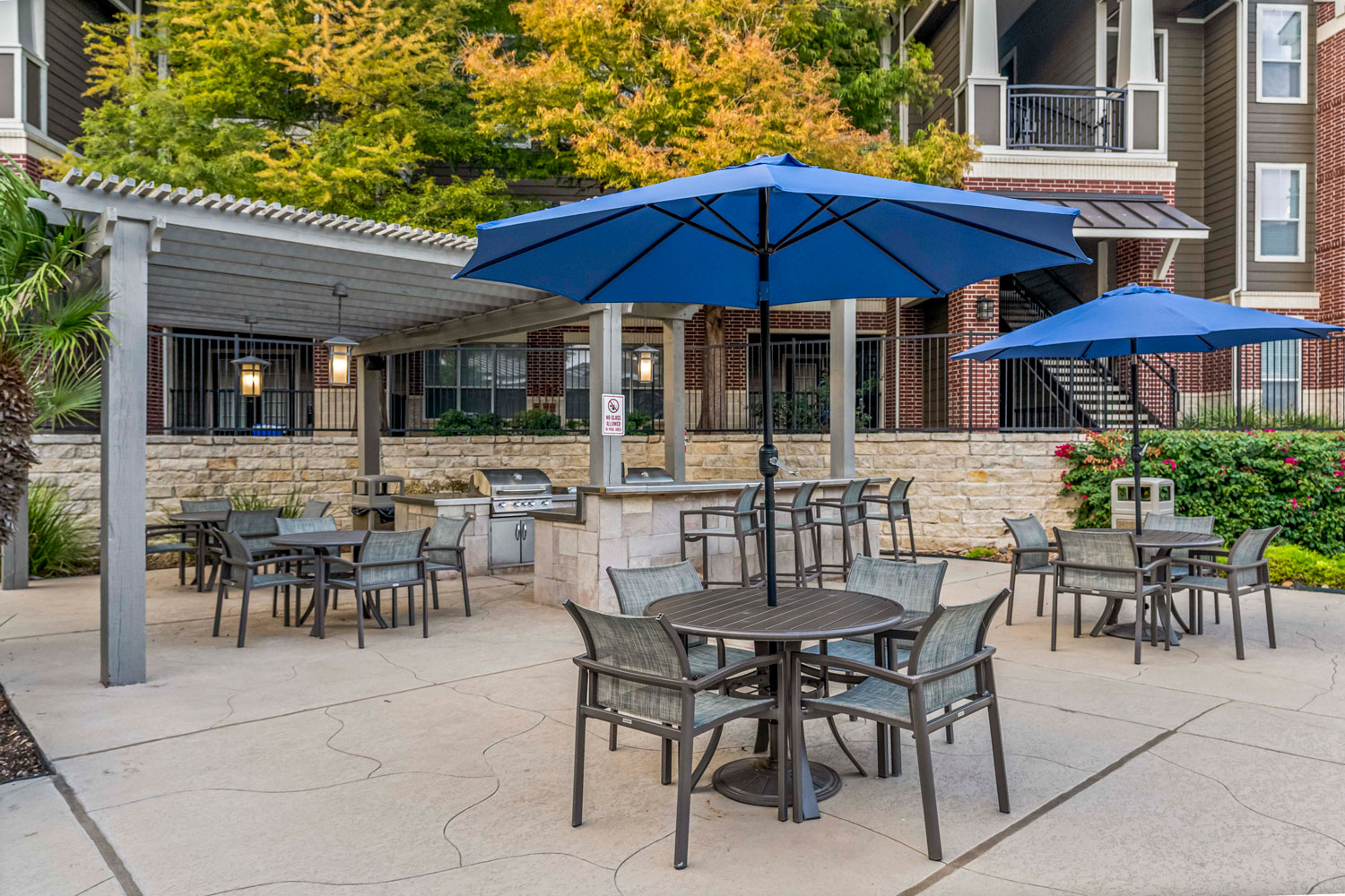 Outdoor grilling pavilion with stainless grills, bar seating, patio dining tables, and blue umbrellas in landscaped apartment courtyard