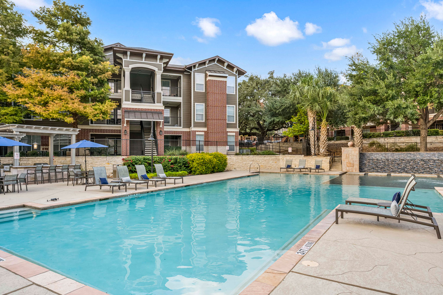Resort style swimming pool with lounge chairs, palm trees, stone water feature wall, and apartment buildings surrounding courtyard