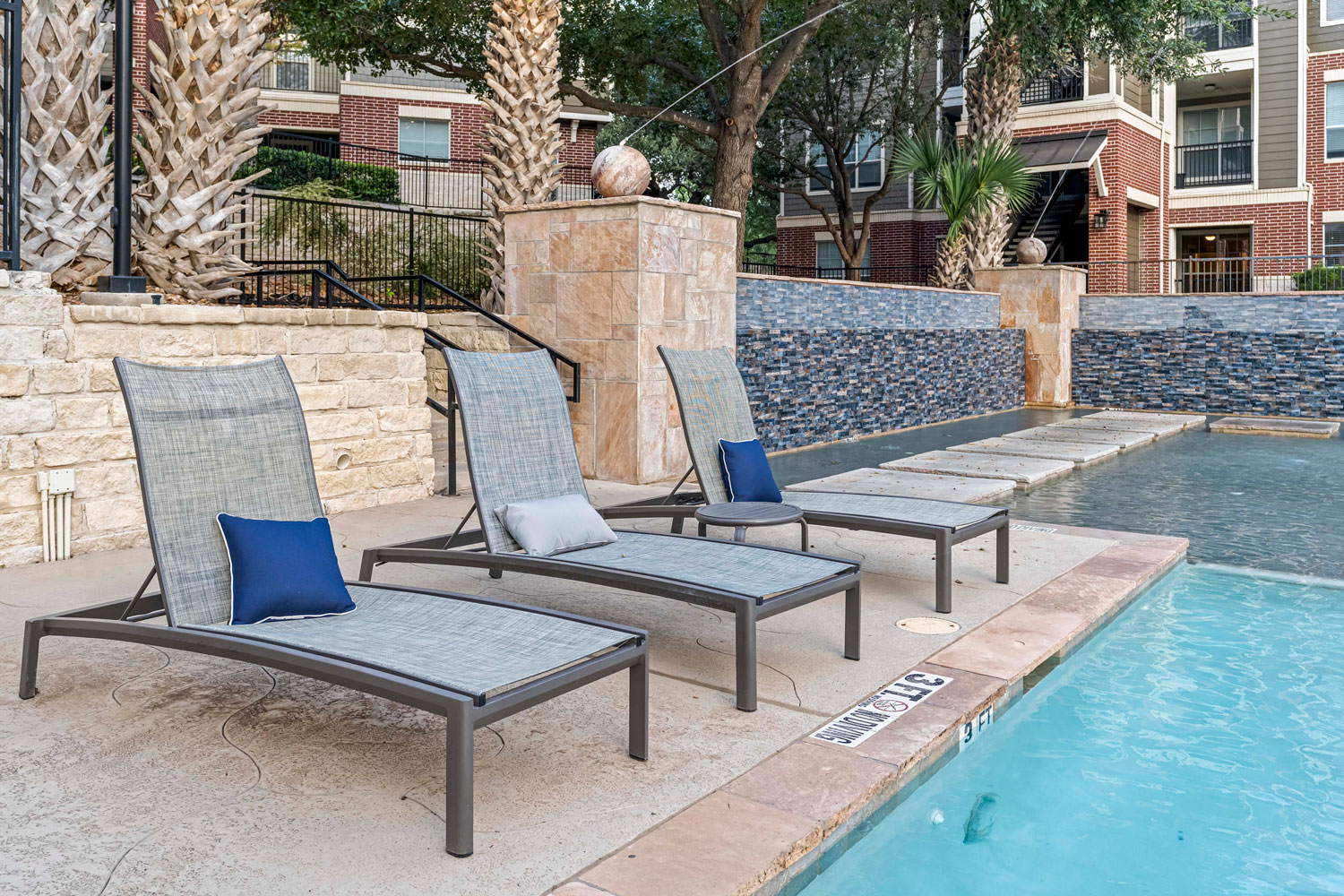 Poolside lounge chairs with cushions beside resort style swimming pool and decorative stone water feature with stepping stones