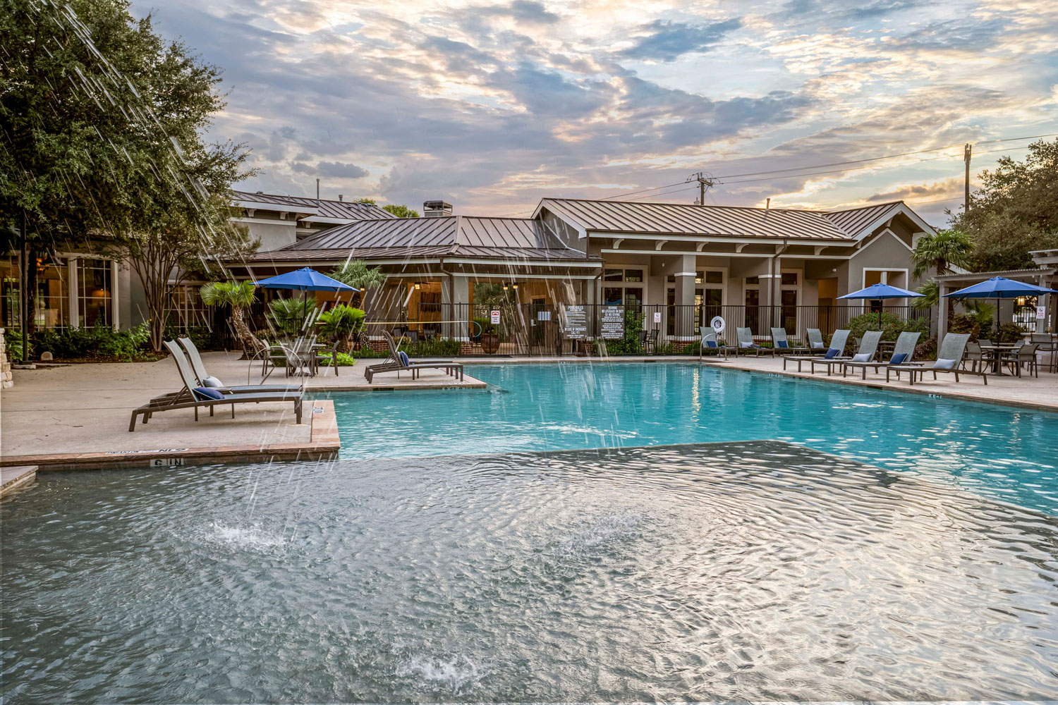 Resort style swimming pool with water fountain feature, lounge chairs, blue umbrellas, and clubhouse building at sunset