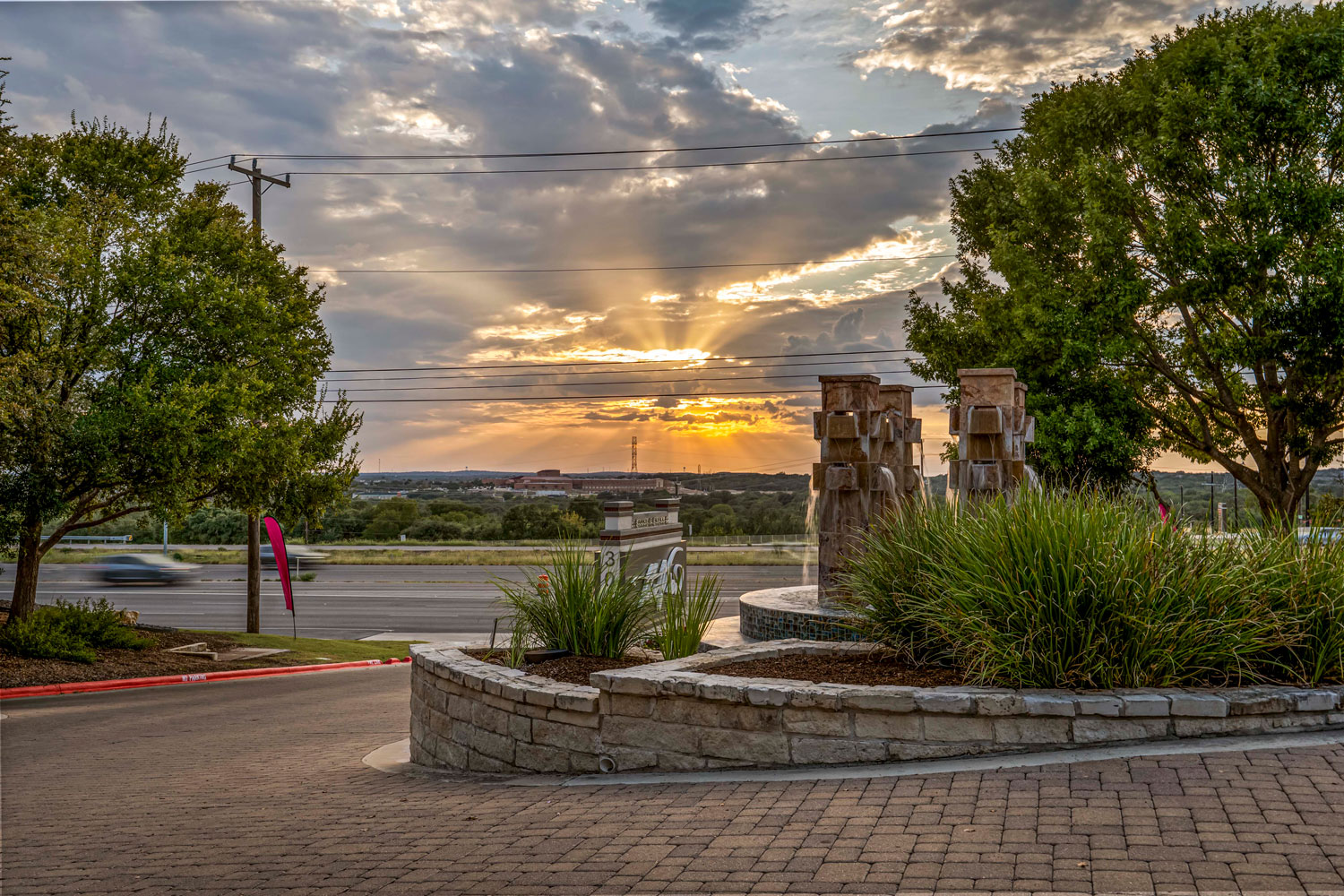 Wide view of the apartment community entrance featuring a stone water fountain, lush landscaping, and a dramatic sunset sky