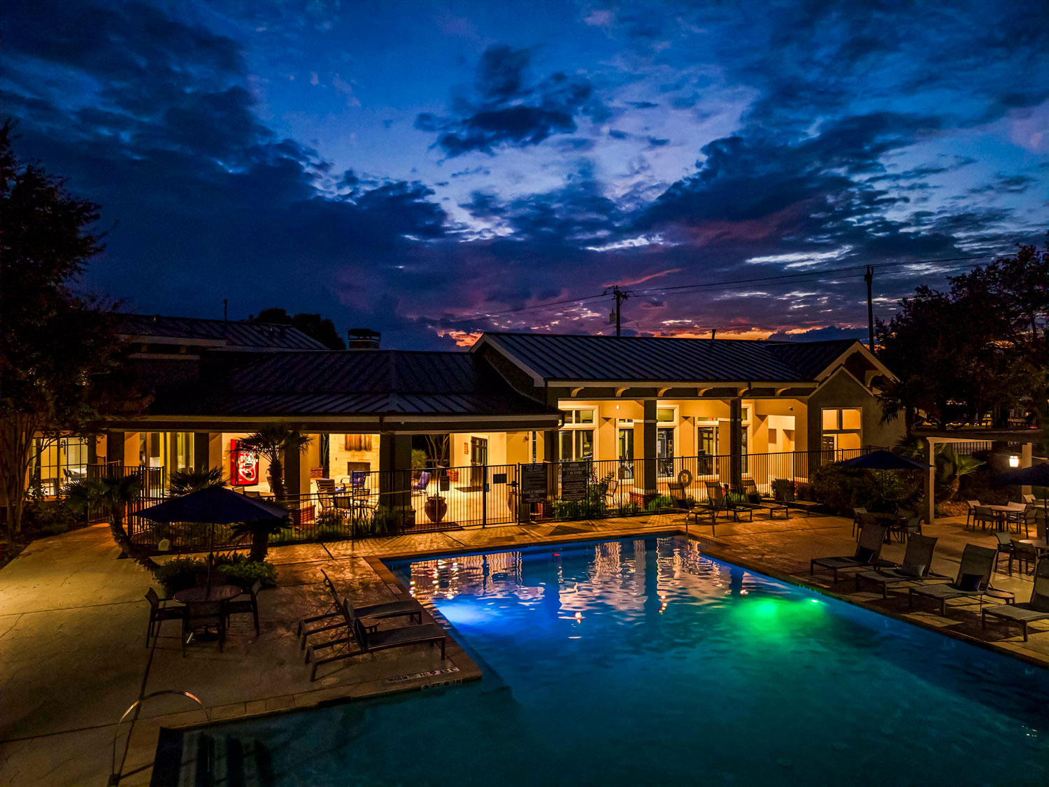 Resort style swimming pool at night with glowing water, lounge chairs, patio seating, and illuminated resident clubhouse