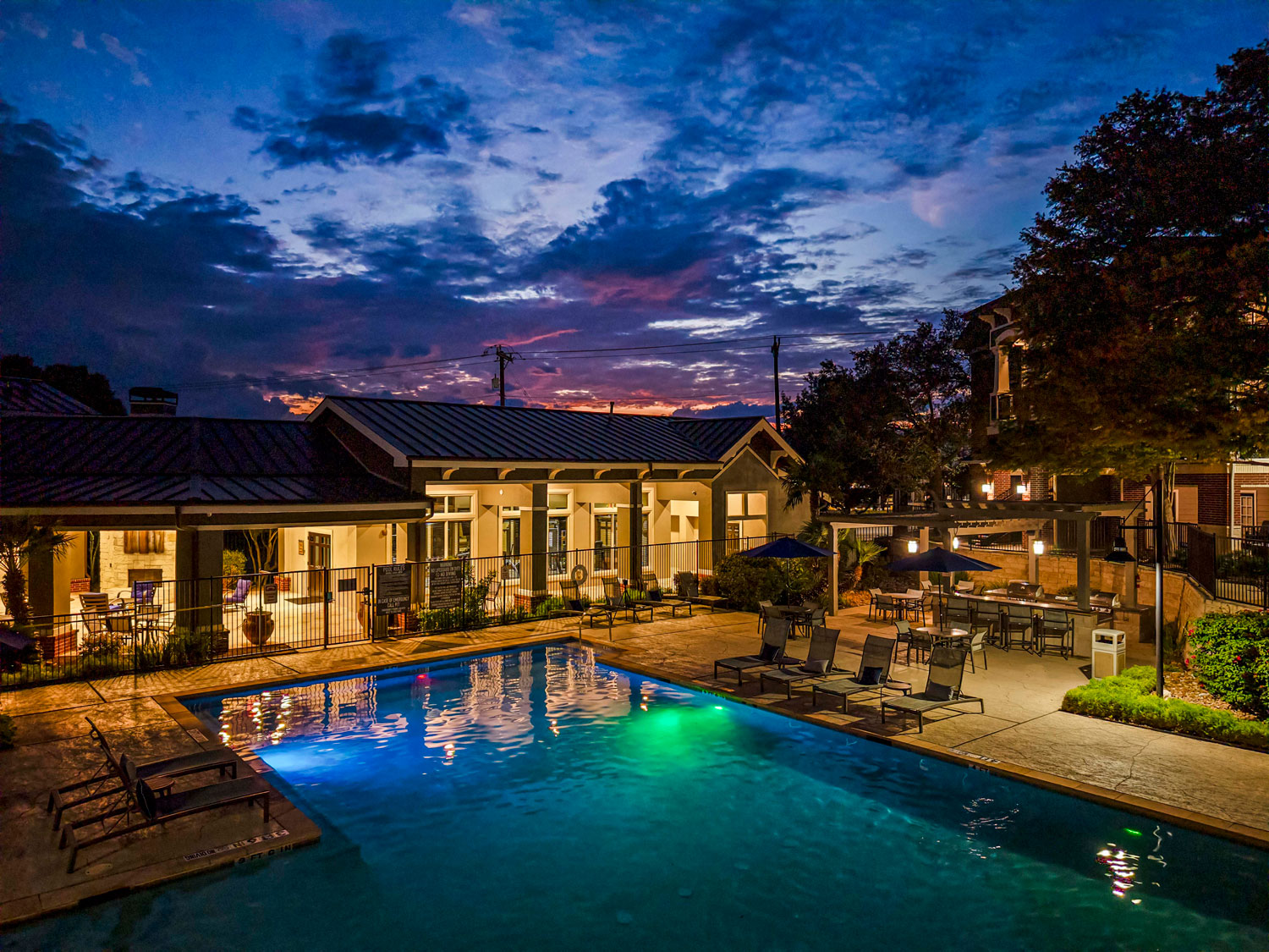 Resort style swimming pool at dusk with illuminated clubhouse, lounge chairs, umbrellas, and landscaped patio seating areas