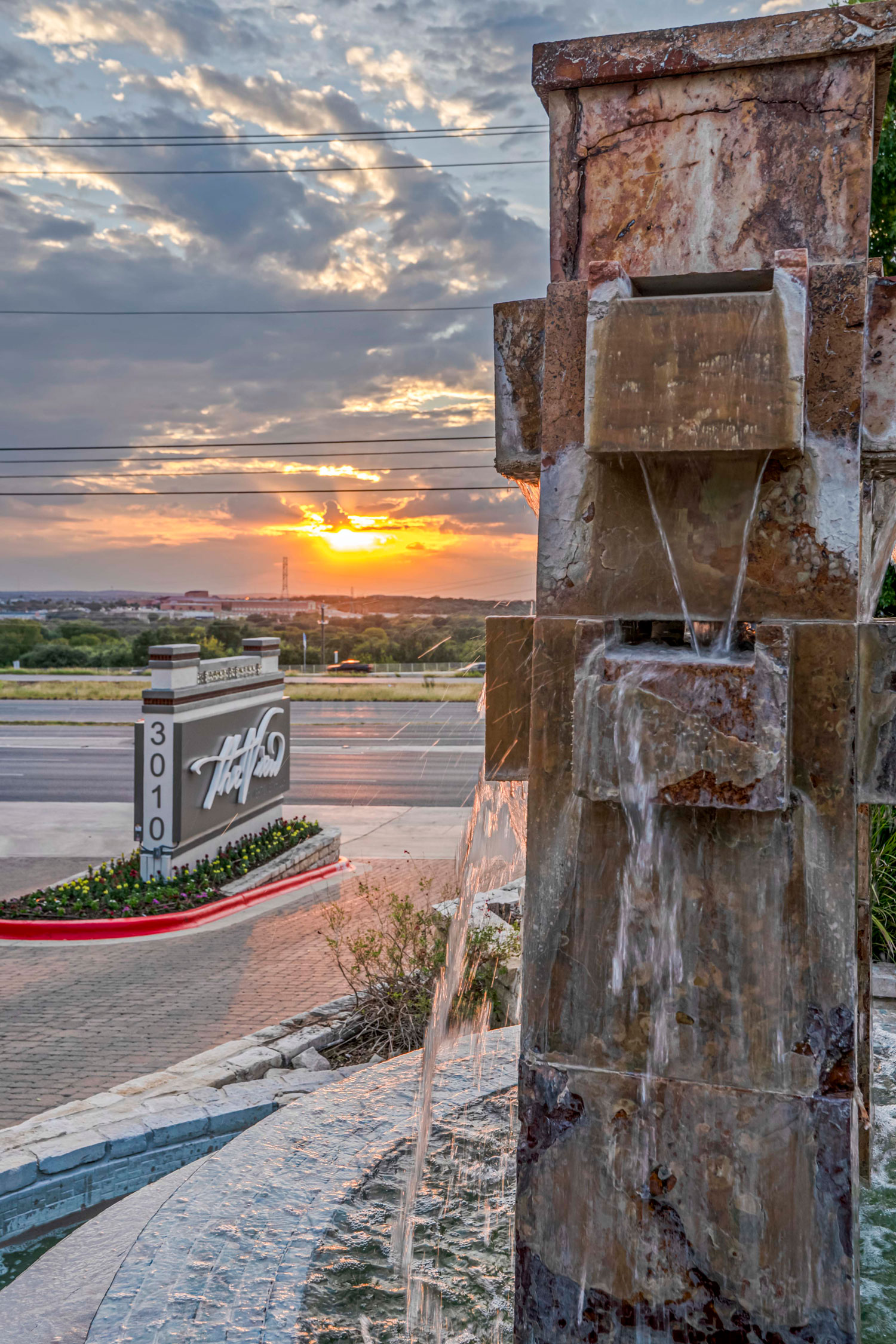 Side view of a stacked stone water fountain feature at sunset with the community entrance sign and brick paver pathway