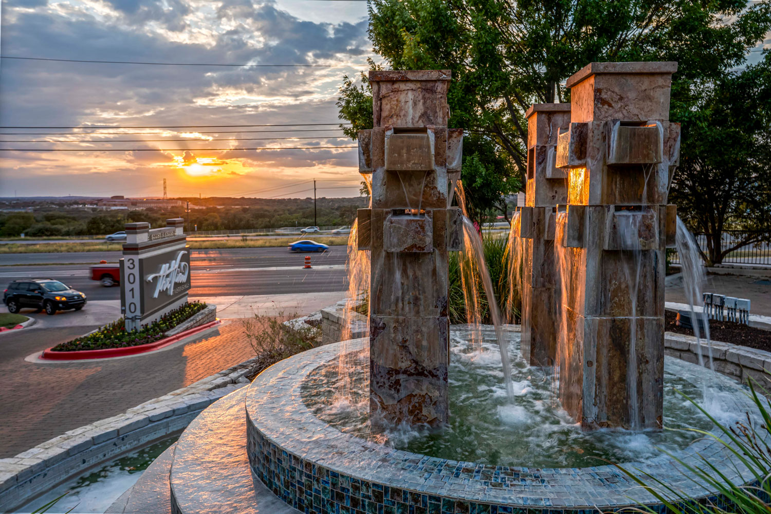 Dual stacked stone water fountain sculptures at the community entrance with a mosaic tile basin and vivid sunset backdrop