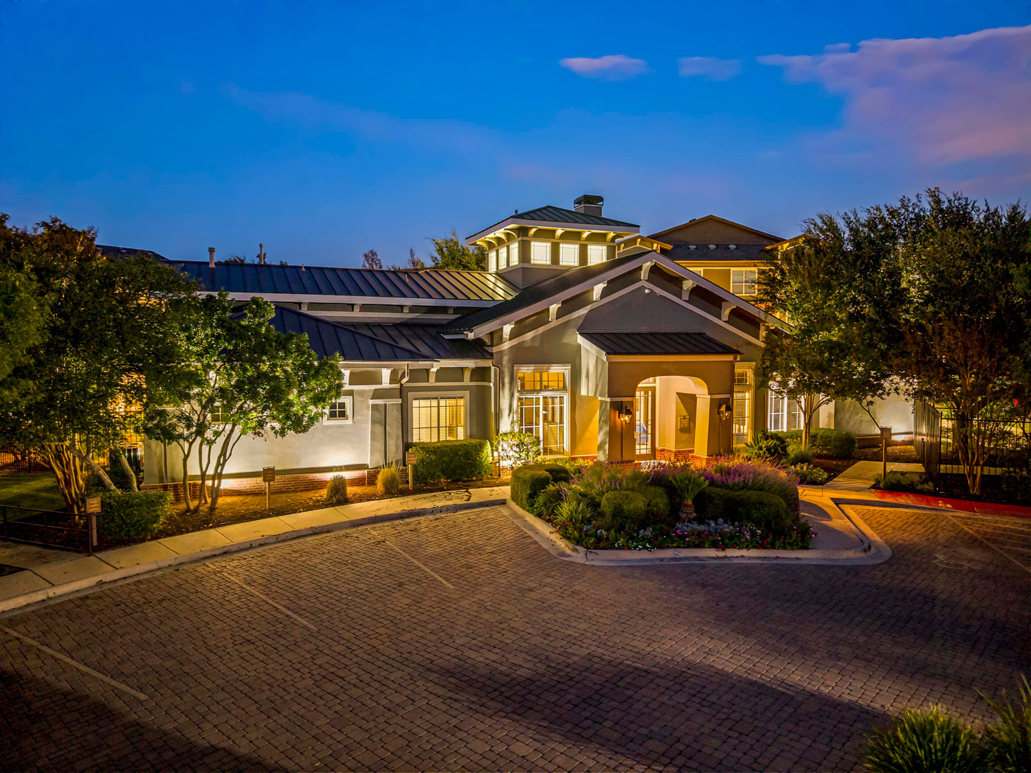 Dusk exterior of an illuminated apartment leasing office with a metal roof, arched entrance, and brick paver driveway