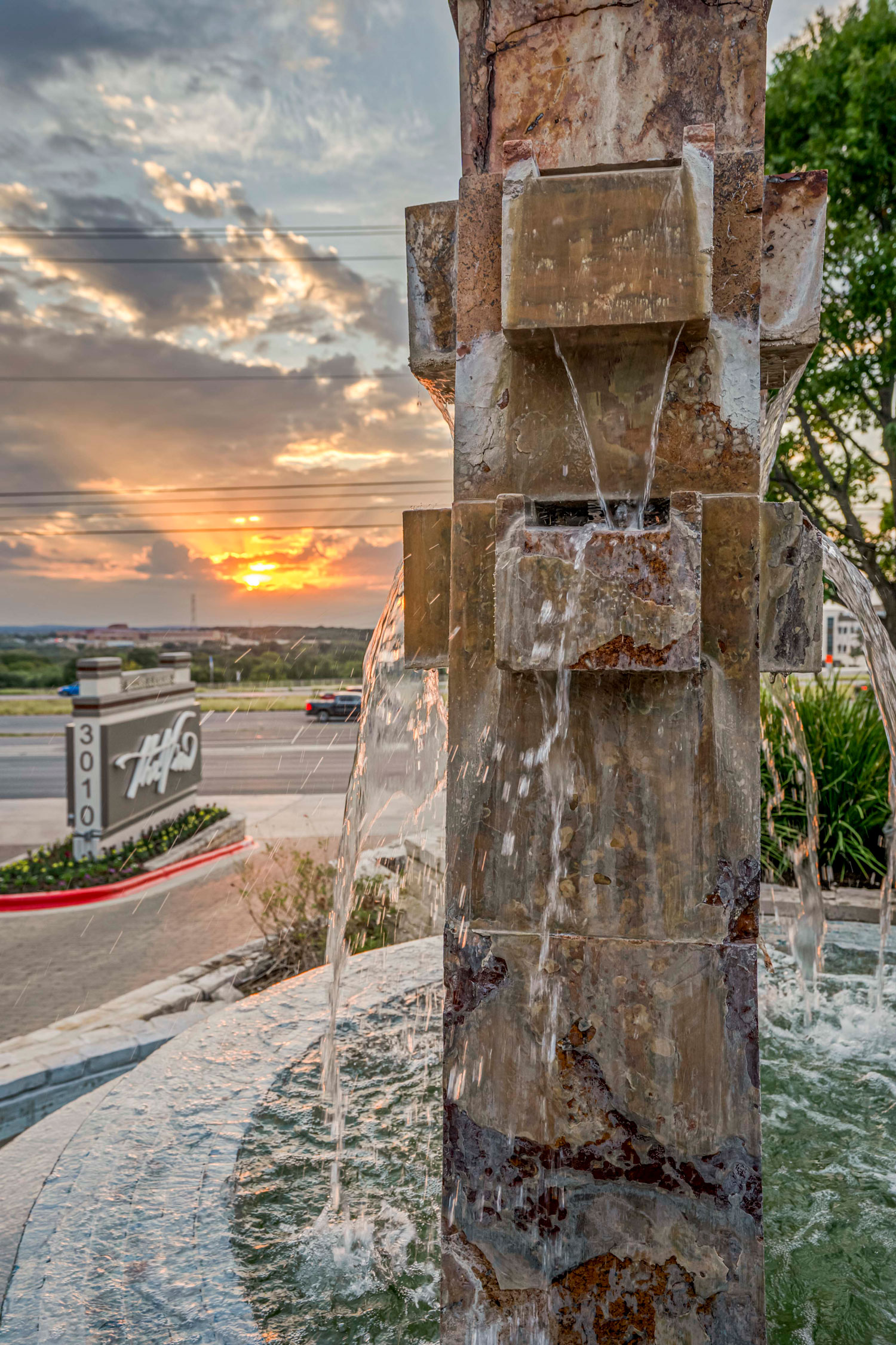 Close-up of a stacked stone water fountain cascading water at sunset with a community entrance sign in the background