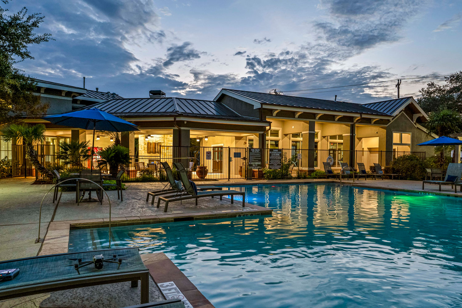 Resort style swimming pool at sunset with lounge chairs, patio seating, blue umbrellas, and illuminated clubhouse in landscaped courtyard