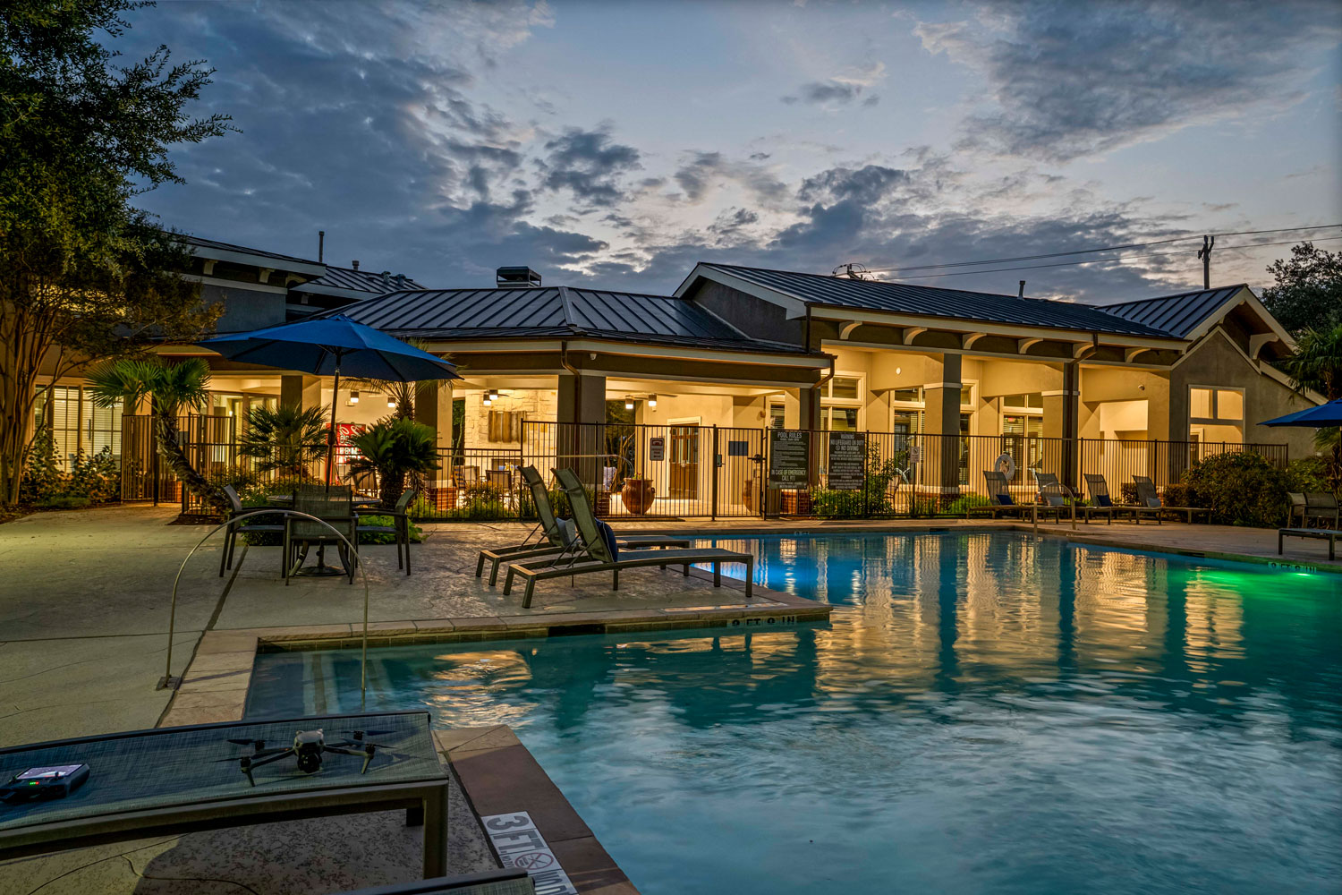 Community clubhouse and pool area at sunset with patio seating, lounge chairs, umbrellas, and softly lit building exterior