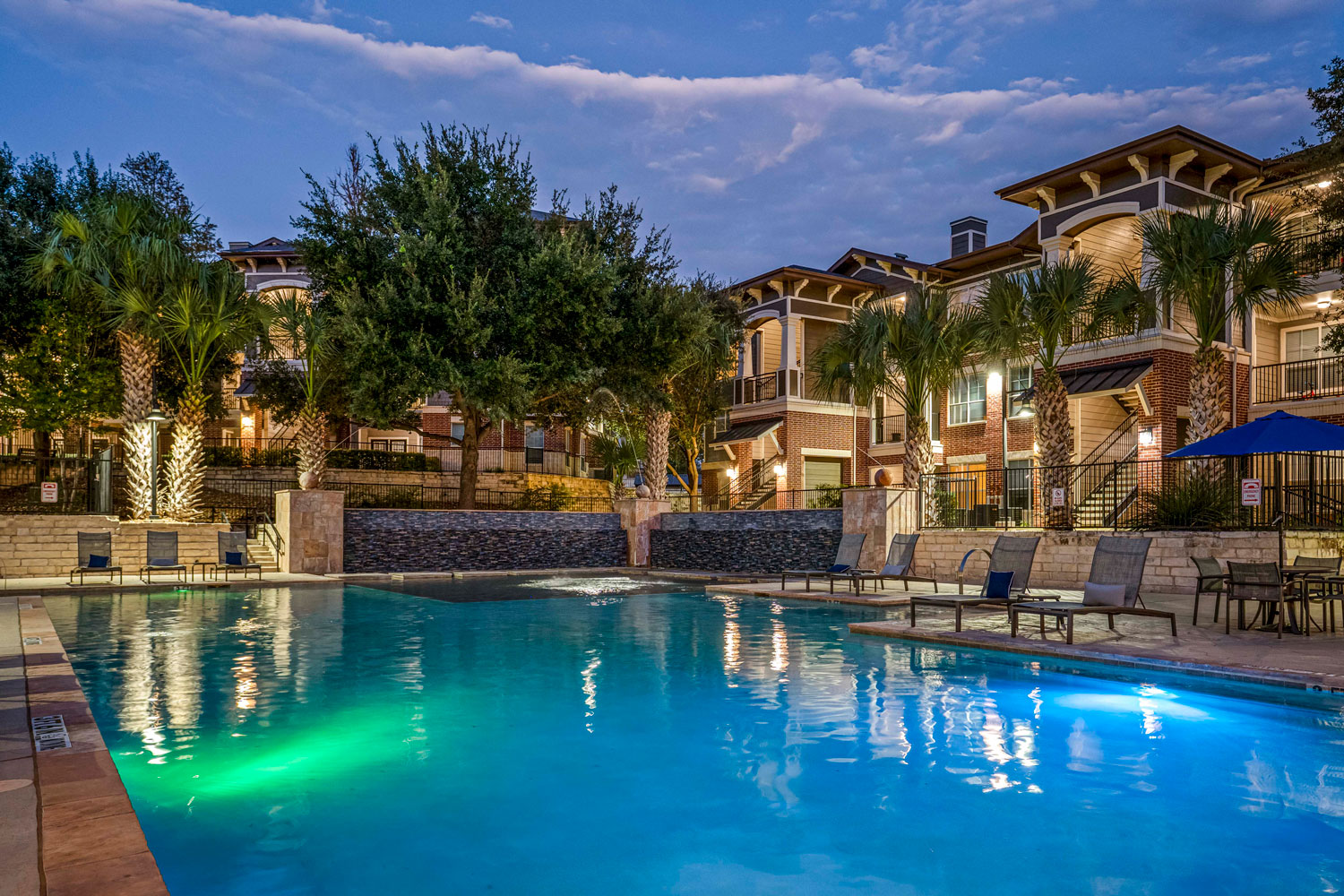 Resort style swimming pool at dusk with illuminated water, lounge chairs, palm trees, and apartment buildings surrounding courtyard