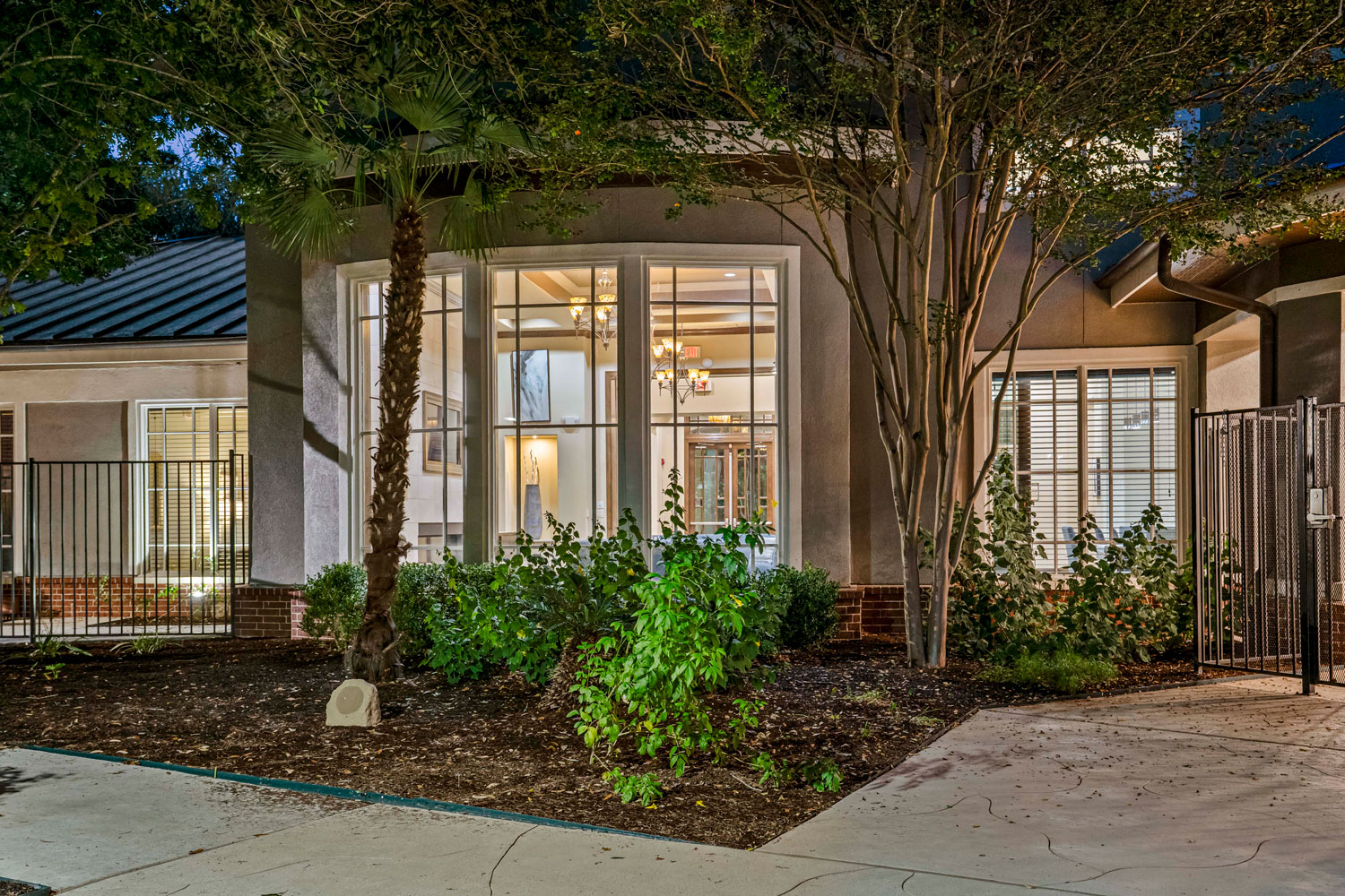Nighttime exterior of a leasing office building with floor-to-ceiling windows, warm interior lighting, and lush landscaping