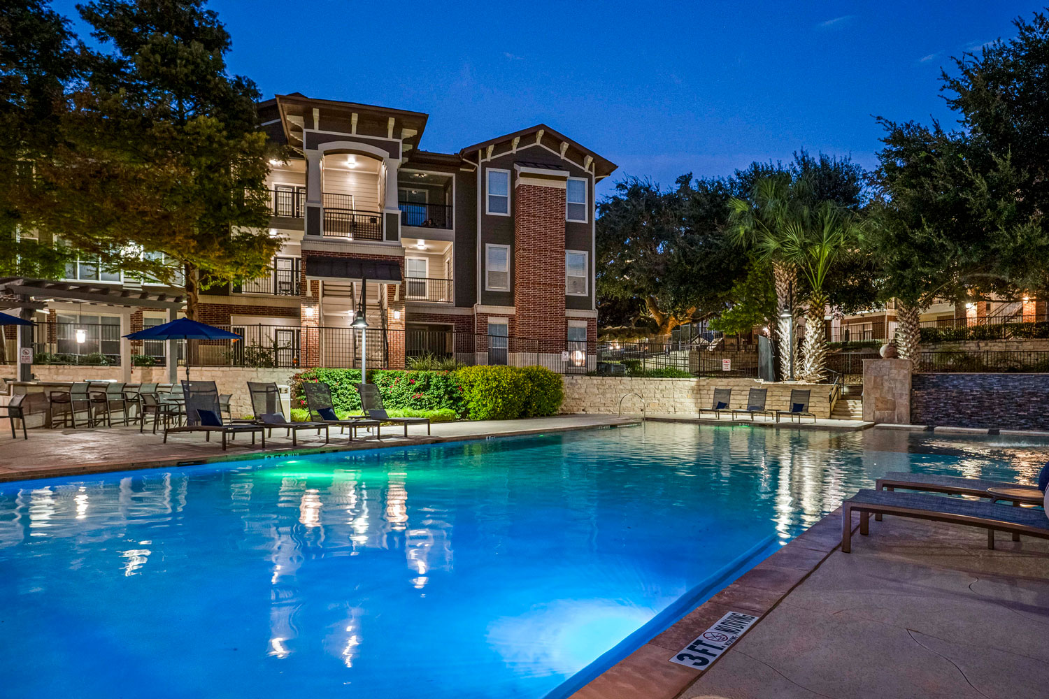 Evening view of resort style swimming pool with glowing water, lounge chairs, patio seating, and illuminated apartment buildings