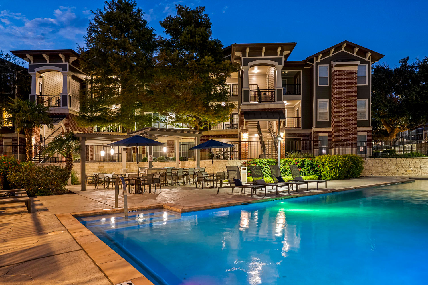 Resort style swimming pool at night with illuminated apartment buildings, lounge chairs, umbrellas, and shaded patio seating