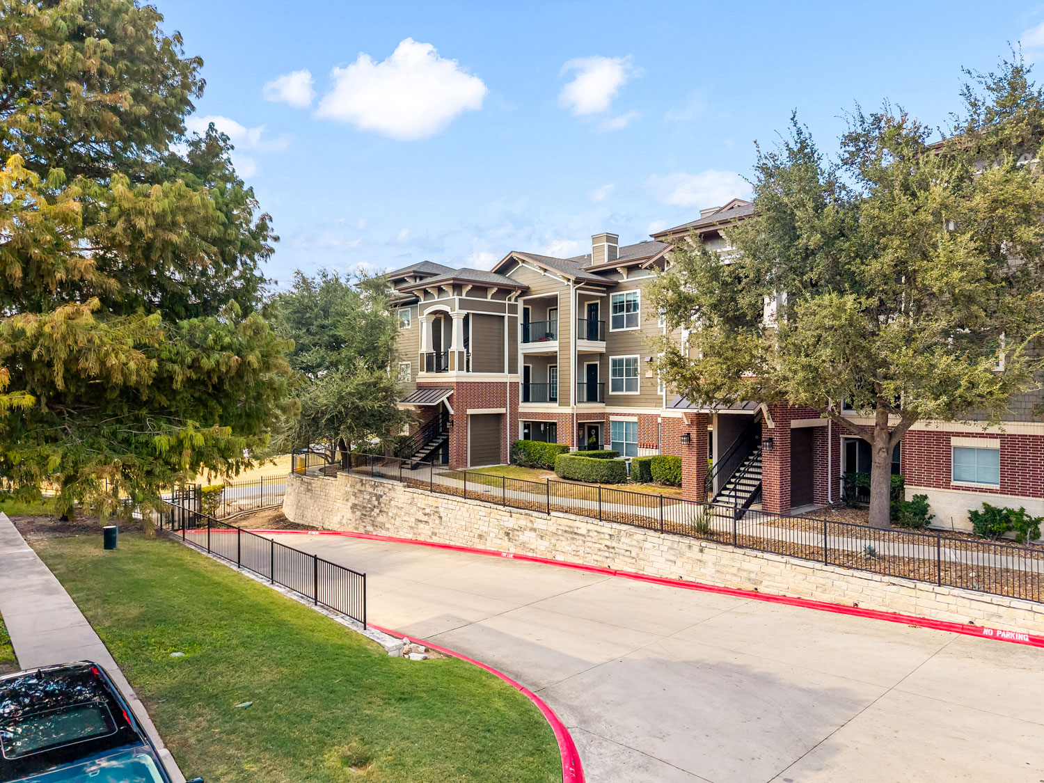 Elevated exterior view of a multi-story brick apartment complex with mature trees, iron fencing, and a wide curved driveway